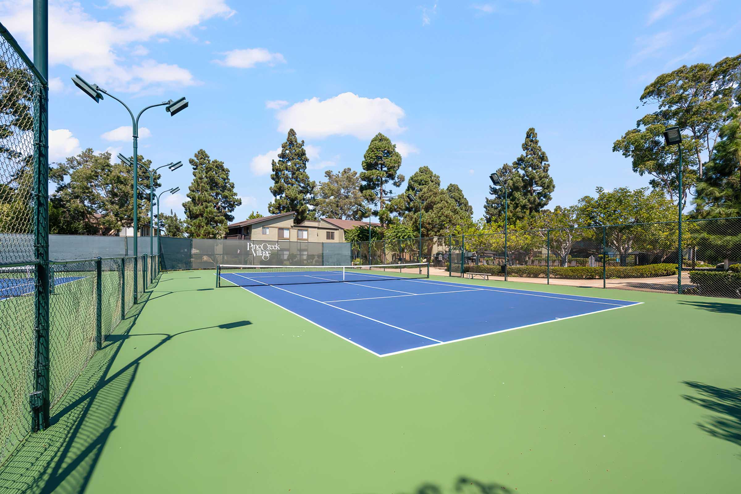 A bright, sunny day at a tennis court featuring two blue tennis courts surrounded by green fencing. Trees line the background, and a building is visible, providing shade. The atmosphere is calm and inviting, ideal for outdoor sports and leisure activities.