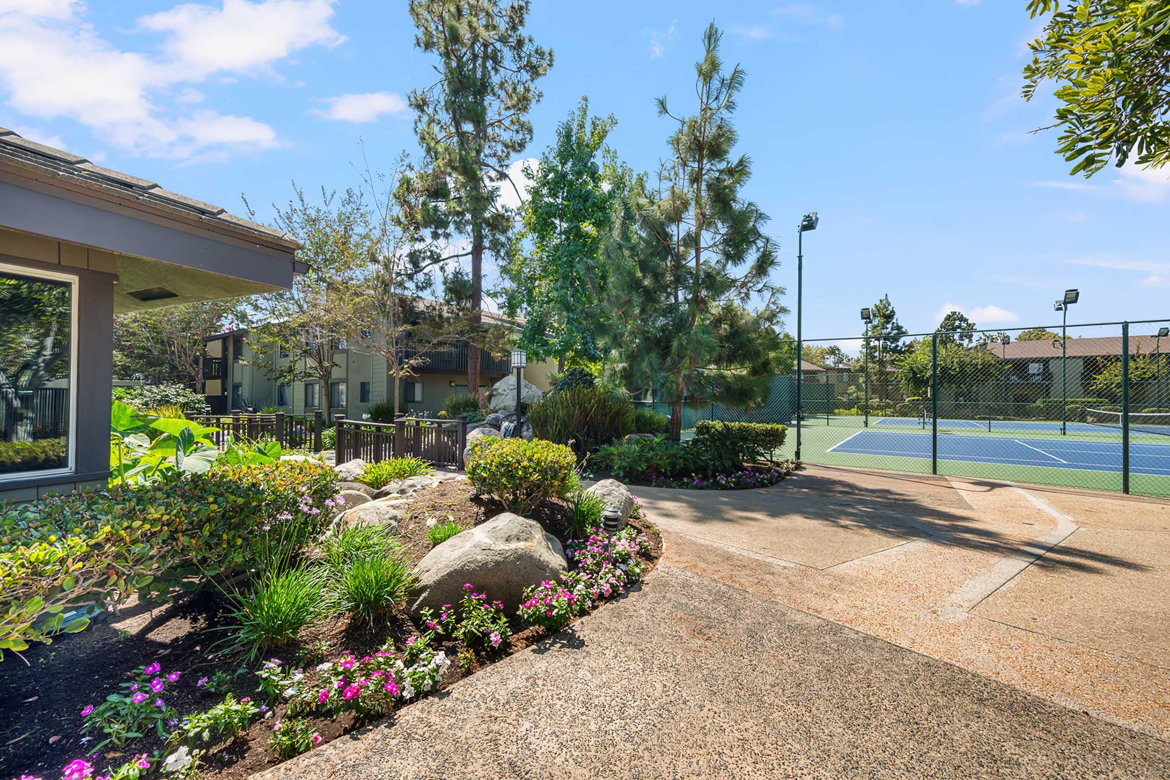 A landscaped path lined with flowers and rocks leads to a tennis court in a residential area. Tall trees and greenery surround the scene, with clear blue skies overhead. The setting is tranquil and well-maintained, showcasing a blend of natural beauty and recreational space.