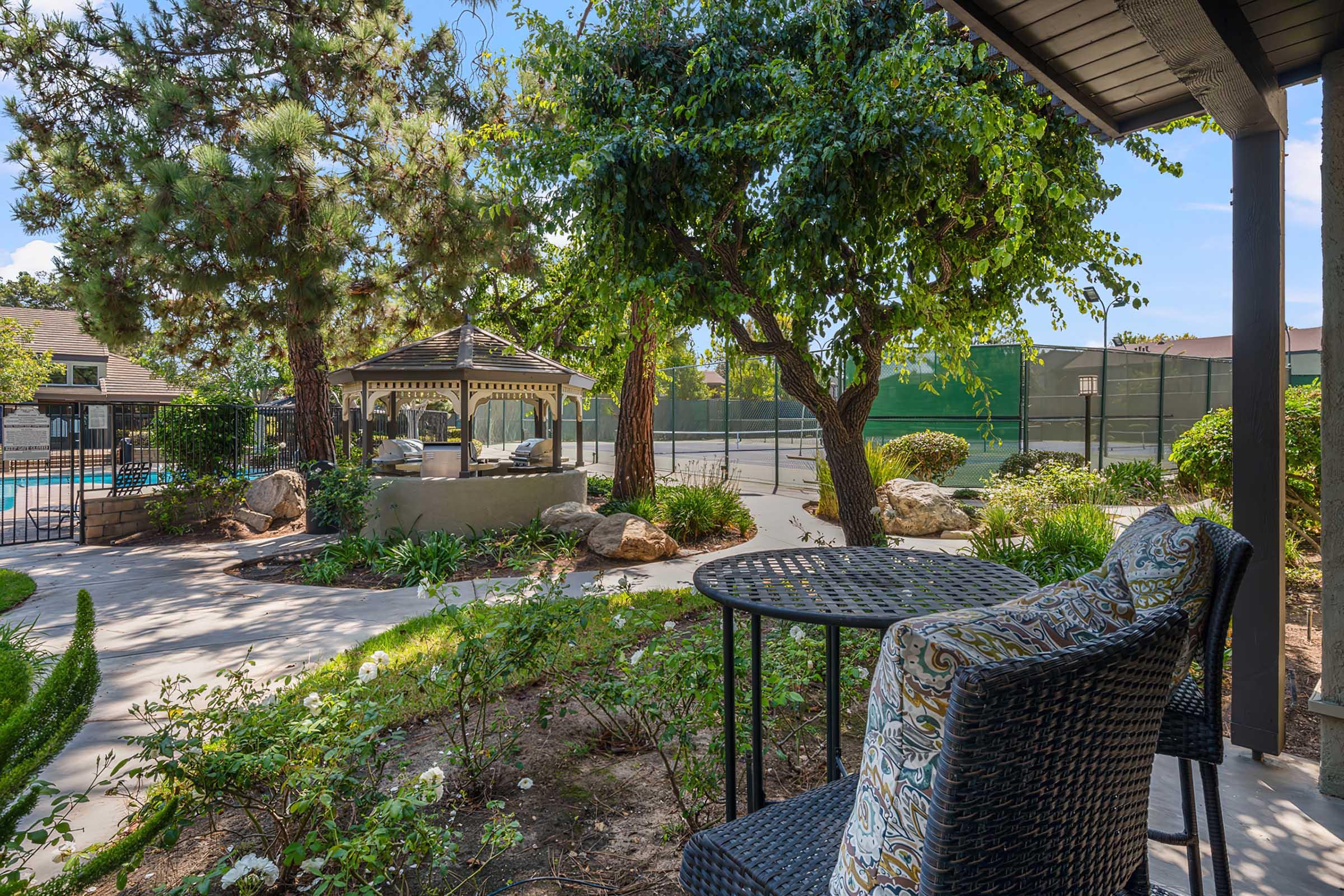 A serene garden view featuring a cozy seating area with a patterned chair beside a small table, surrounded by lush greenery. In the background, a gazebo is visible alongside a swimming pool and tennis courts, creating a peaceful outdoor retreat.