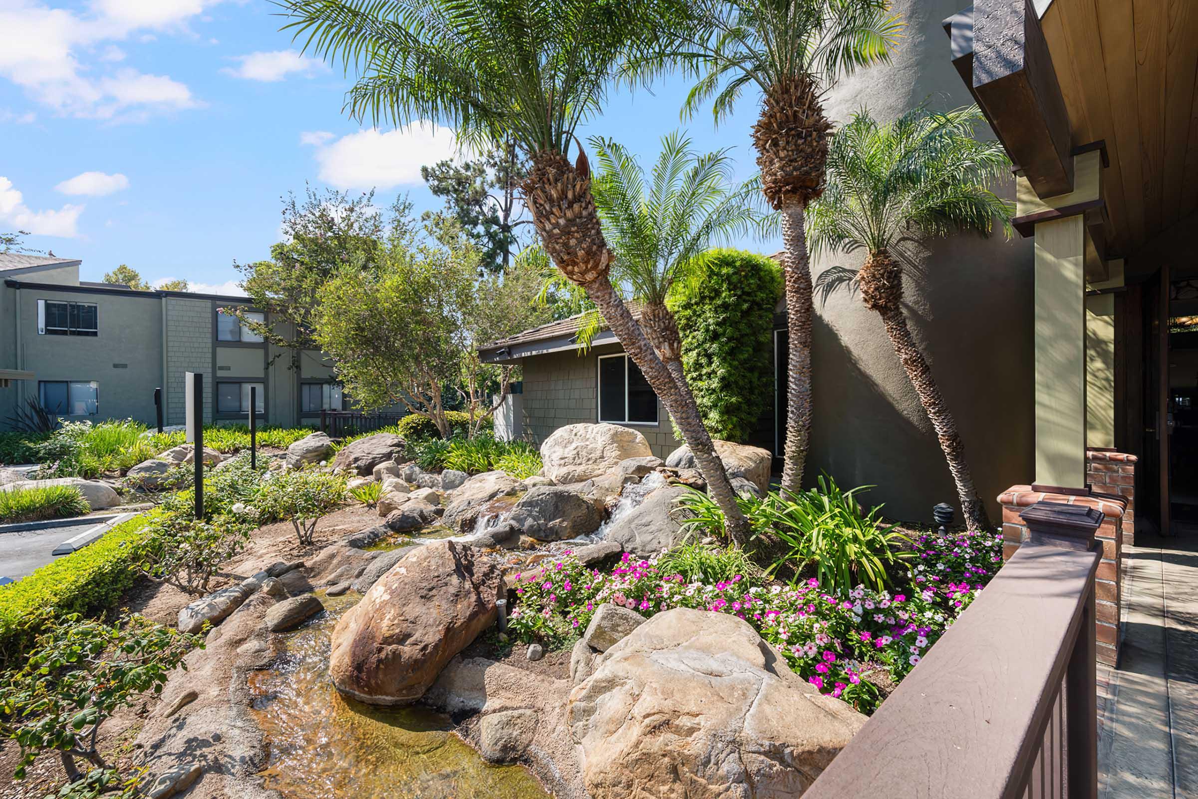 Lush landscaping featuring palm trees, flowering plants, and large rocks near a building. A small water feature adds to the serene atmosphere, with a pathway leading through the garden. Sunlight shines on the greenery, creating a vibrant and inviting outdoor space.