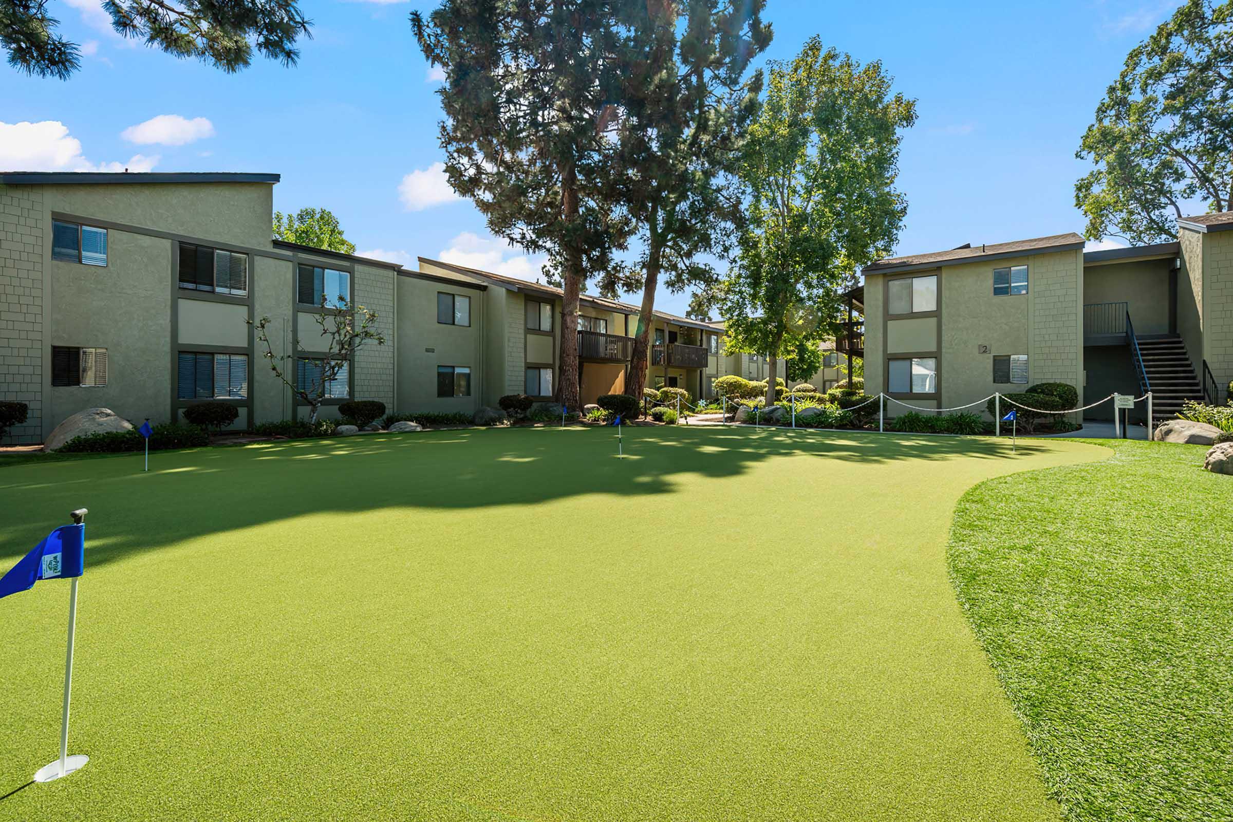 Lush green lawn with a small putting green marked by a blue flag in the foreground. In the background, there are two-story apartment buildings surrounded by trees and shrubs, under a clear blue sky.