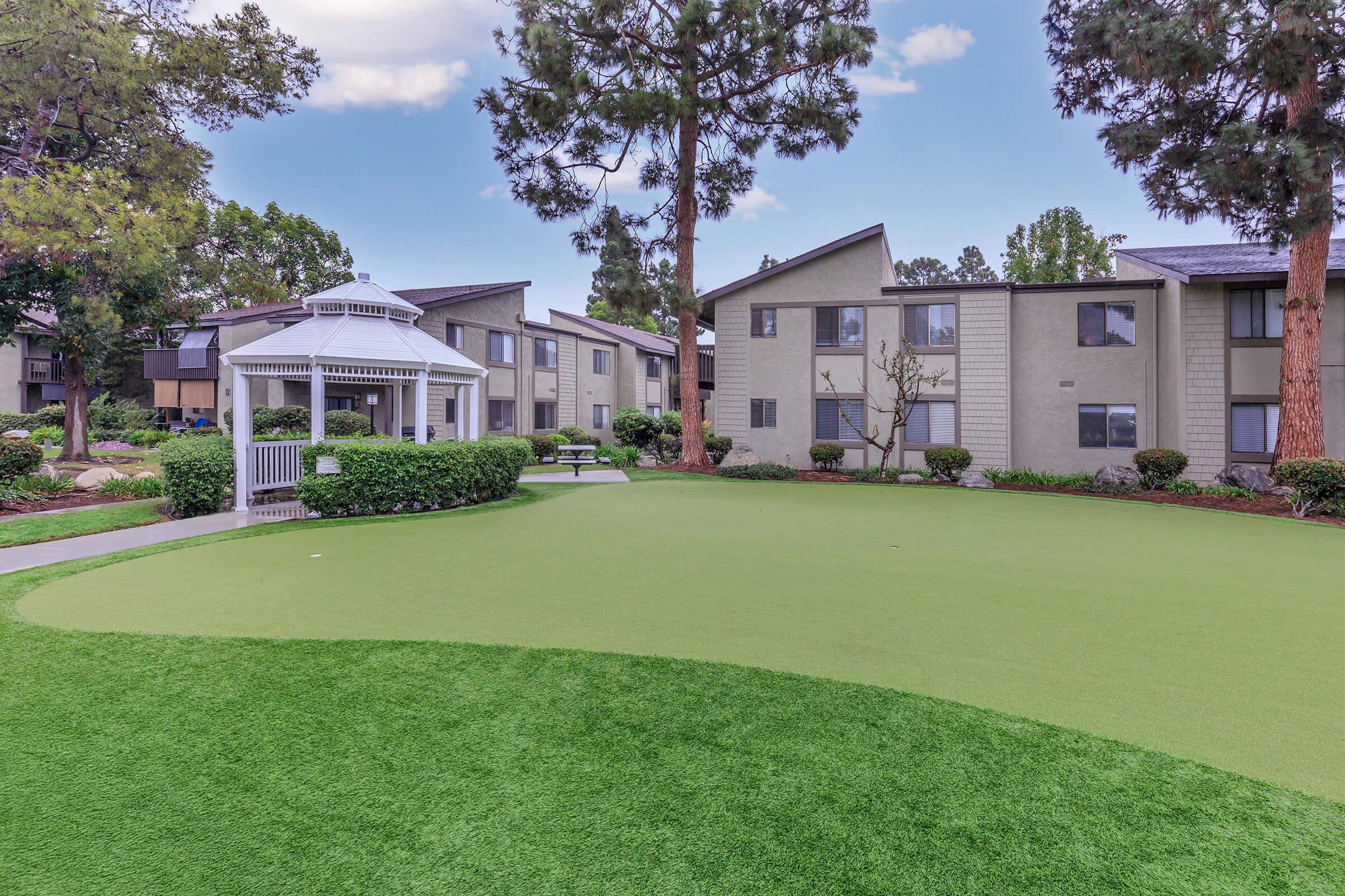 A landscaped recreational area featuring a lush green lawn, a gazebo, and apartment buildings in the background. The scene is framed by trees and well-maintained shrubs, creating a serene outdoor space for residents to enjoy.