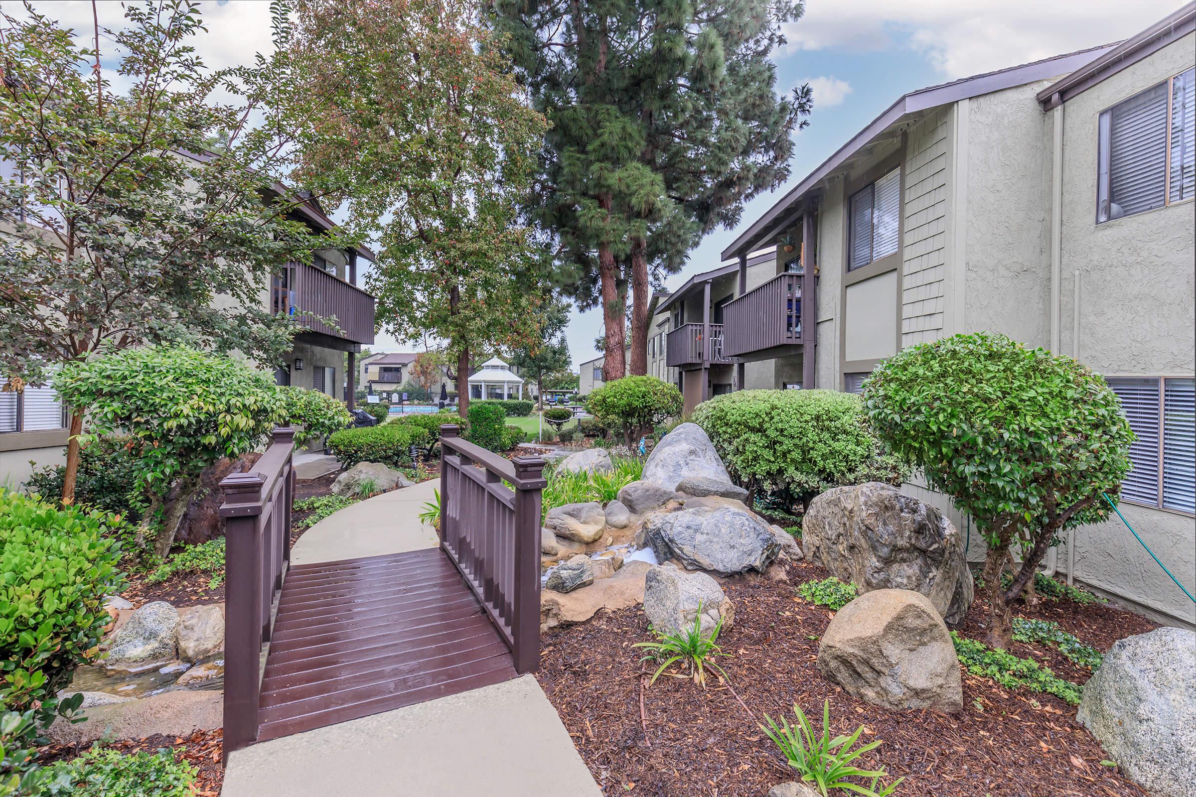 A peaceful residential pathway in an apartment complex featuring a wooden bridge over a small stream, surrounded by manicured greenery, rocks, and trees. The scene conveys a tranquil atmosphere with well-maintained landscaping and nearby buildings visible.