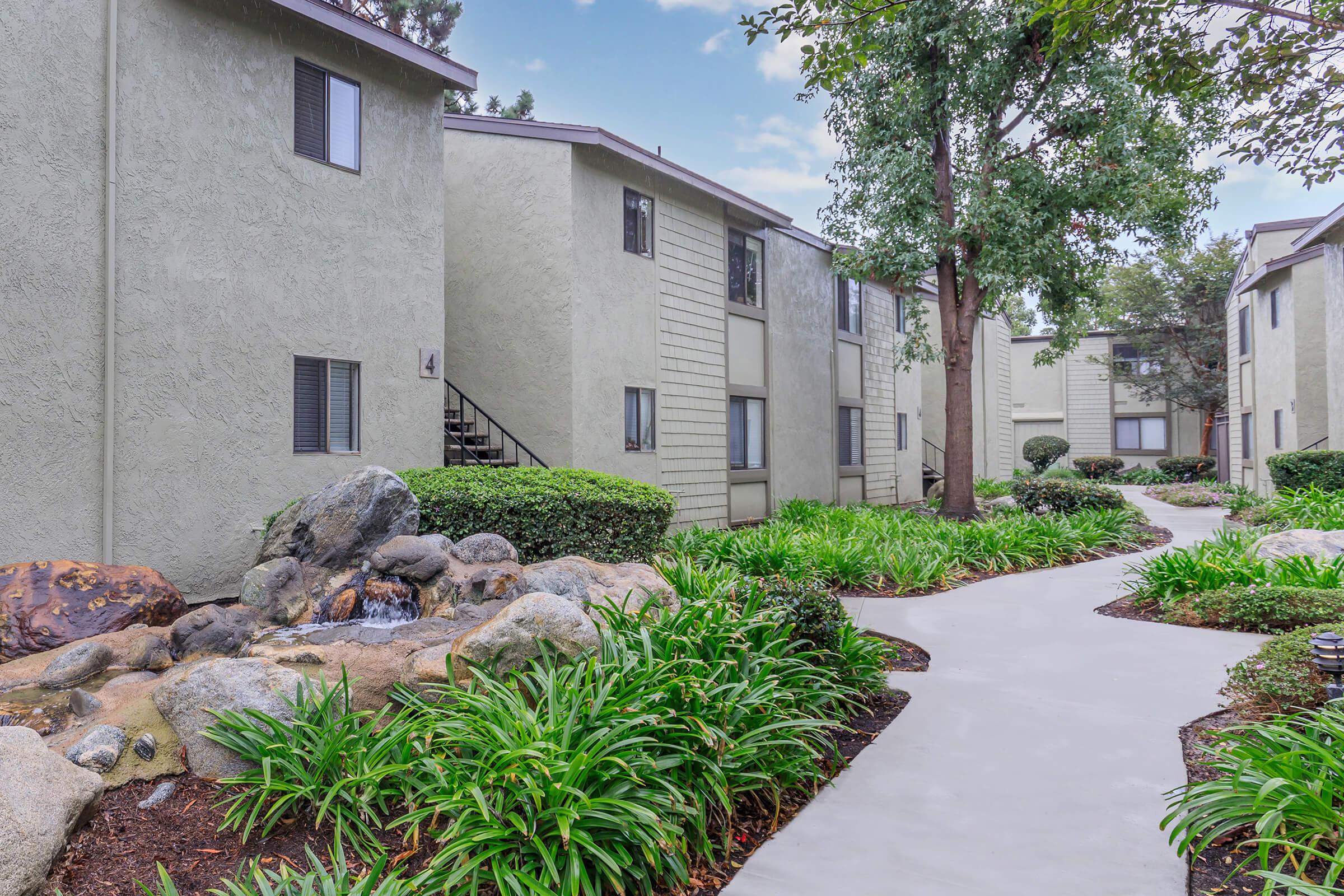 A well-maintained apartment complex featuring two-story buildings surrounded by lush greenery. A winding walkway leads through neatly trimmed shrubs and decorative rocks, with a small water feature visible, enhancing the peaceful atmosphere. Trees provide shade along the path.