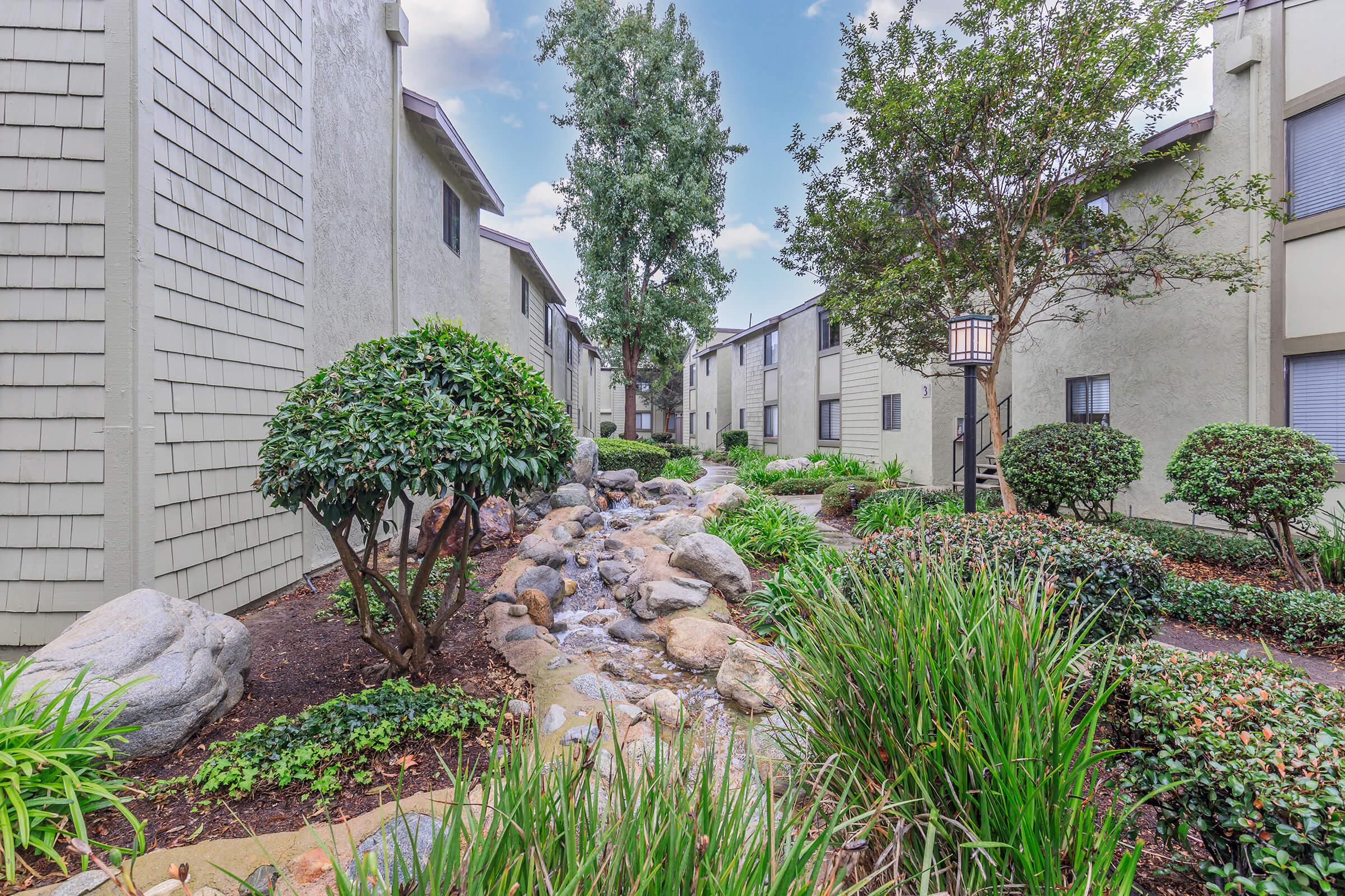 Lush green courtyard featuring a small rock-lined stream flowing through between apartment buildings. The area is surrounded by manicured shrubs and trees, with a lantern-style light fixture visible. The sky is partly cloudy, creating a serene atmosphere.
