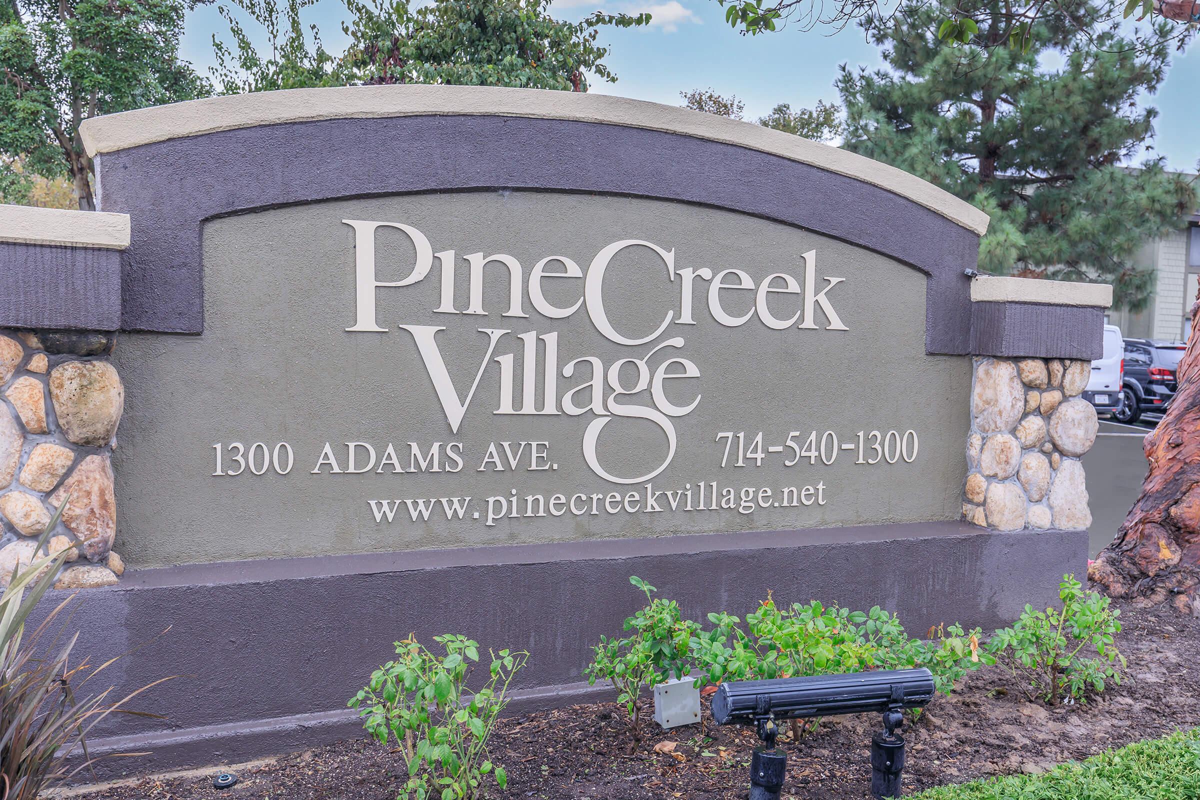 Sign for Pine Creek Village displaying the address "1300 Adams Ave," phone number "714-540-1300," and website "www.pinecreekvillage.net." The sign features a decorative stone base and is set against greenery and landscaping.
