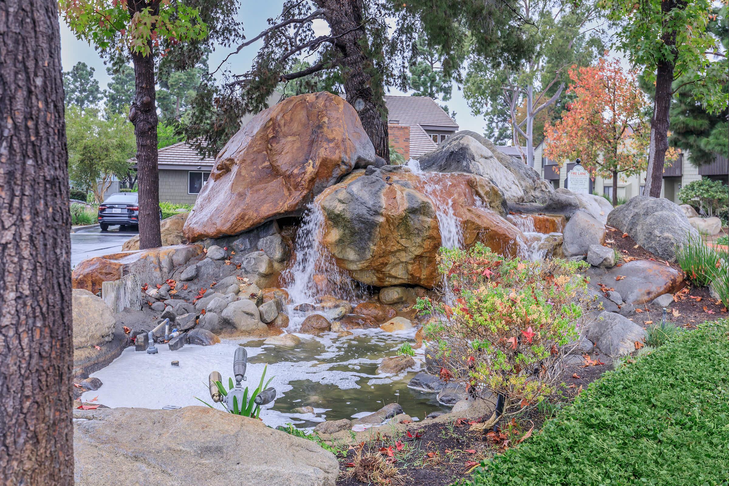 A serene landscape featuring a small waterfall cascading over large rocks into a pond. Surrounding the water feature are lush green plants and colorful autumn foliage. In the background, there are residential buildings partially obscured by trees. The scene conveys relaxation and natural beauty.
