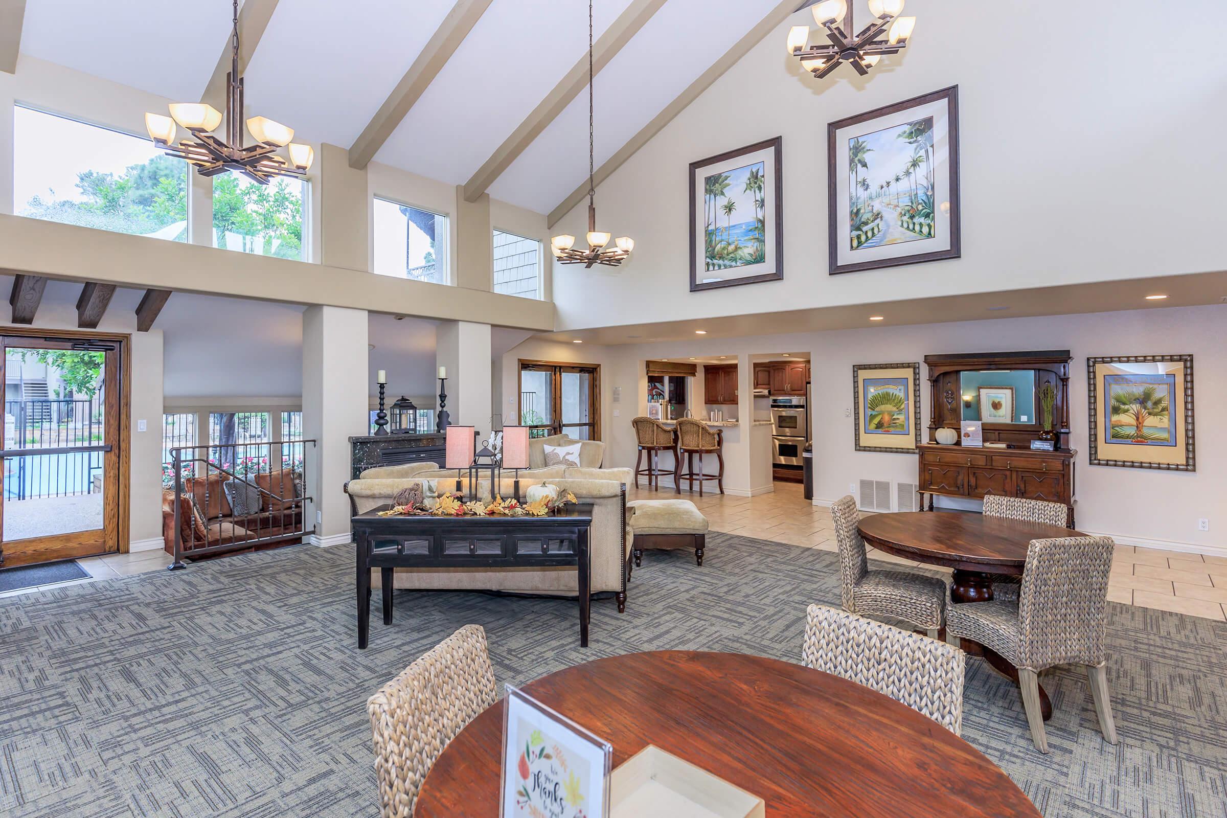 Spacious living area with high ceilings, large windows, and stylish decor. Features comfortable seating, a dining table, and wall art depicting landscapes. A kitchenette is visible in the background, and a cozy entryway leads outside, creating an inviting atmosphere. Natural light fills the space.