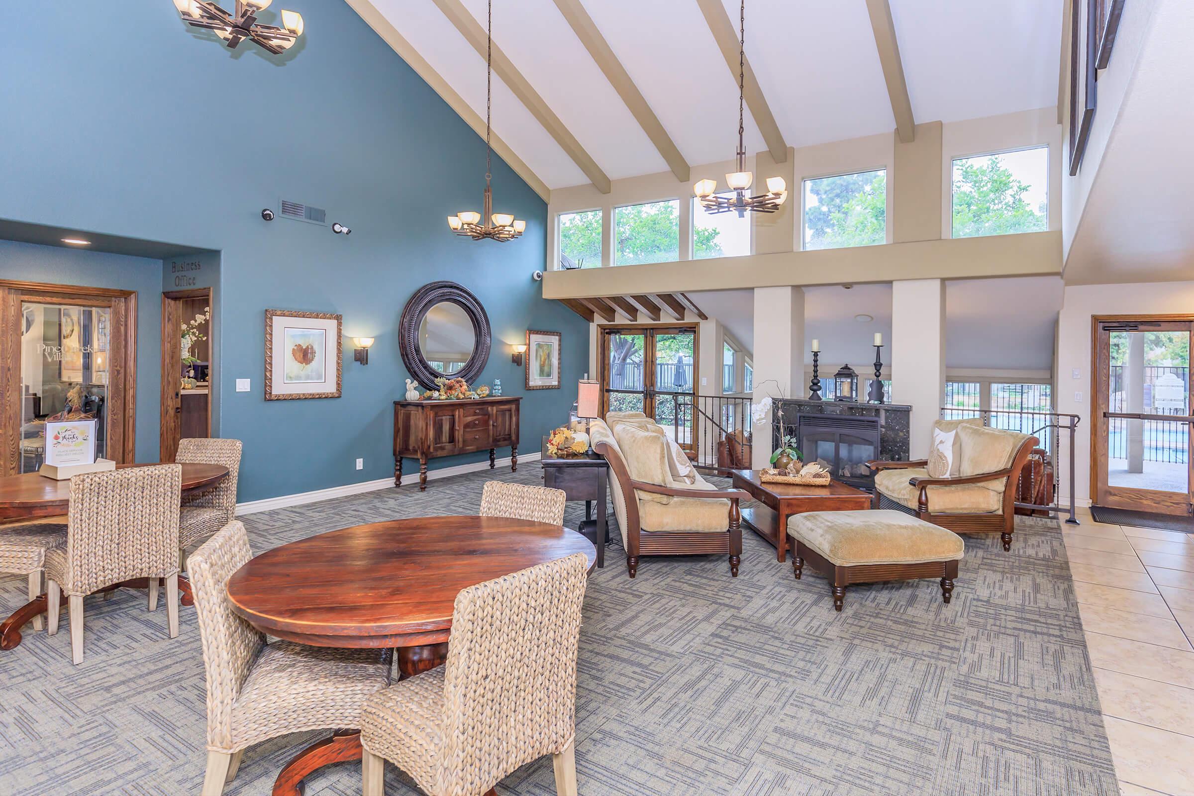 Interior of a spacious lobby with high ceilings, featuring a mix of comfortable seating, decorative tables, and large windows letting in natural light. The walls are painted in a soft blue, and there are decorative elements like artwork and plants, creating a welcoming atmosphere.