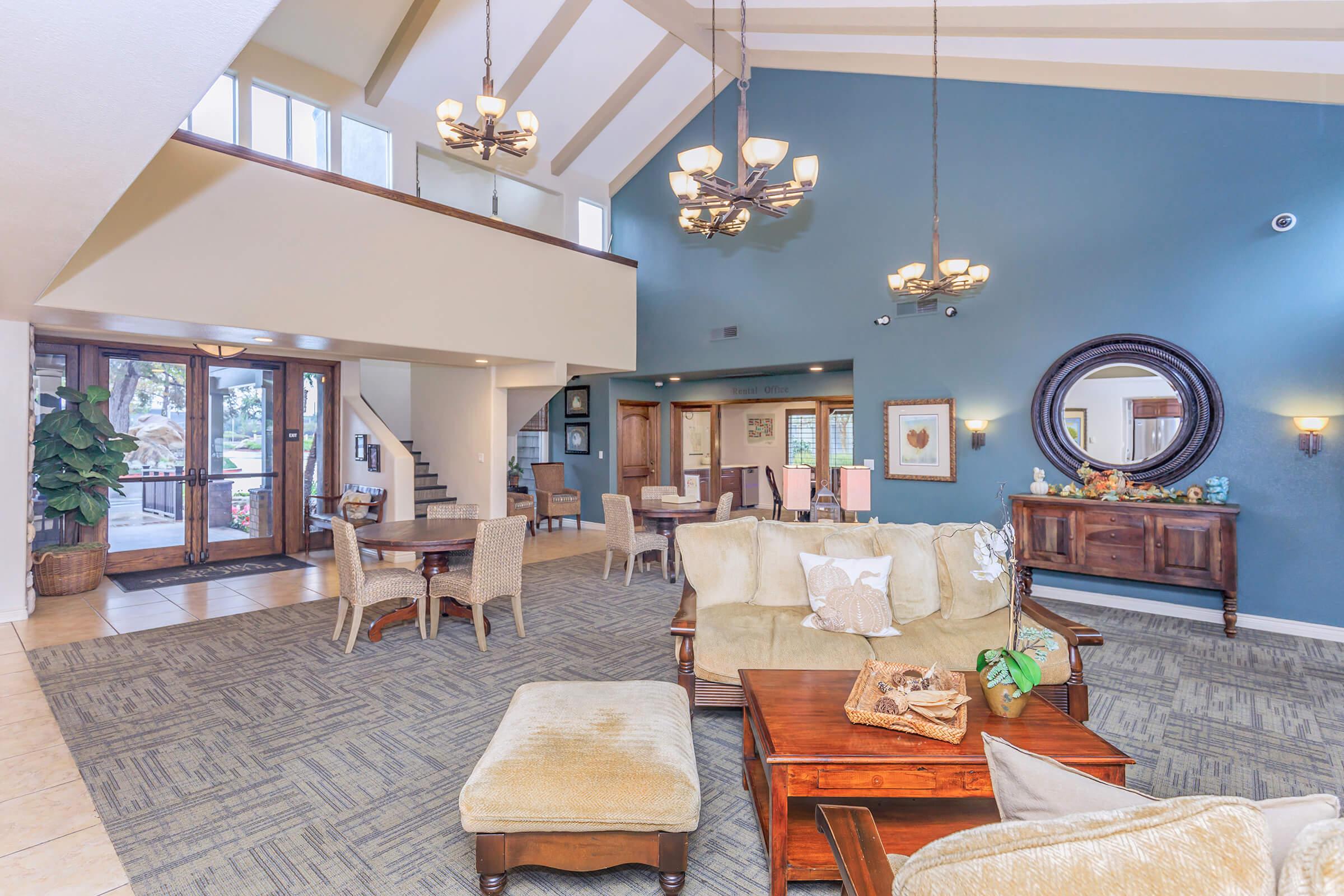 A spacious, elegantly designed lounge area featuring high ceilings with beams, large windows, and chandeliers. There are comfortable sofas and chairs, wooden furniture, and a decorative round mirror. A plant adds a touch of greenery, and the area is well-lit, creating a welcoming atmosphere.