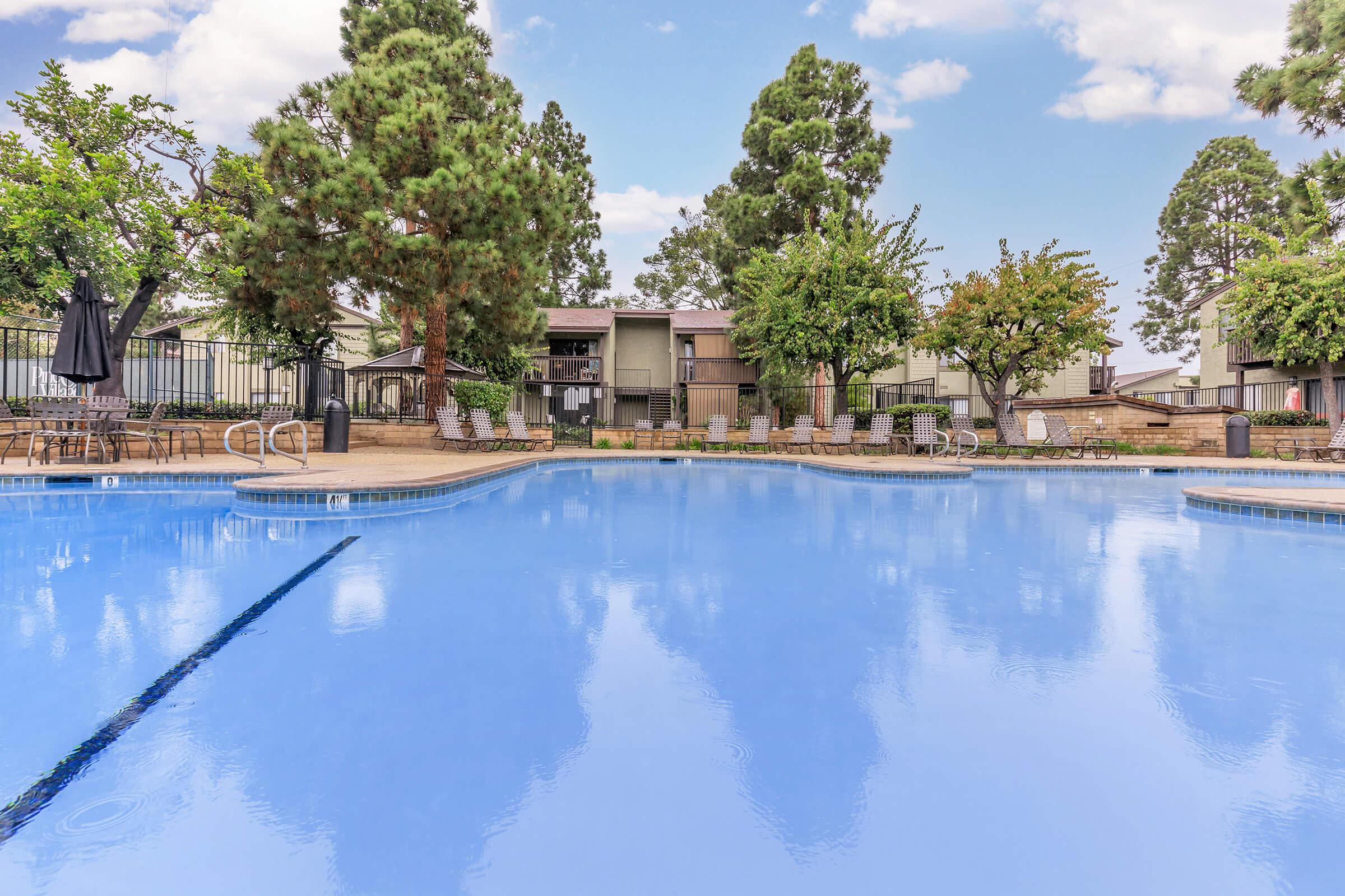 A clear blue swimming pool surrounded by trees and landscaped greenery, with lounge chairs arranged around the pool area. In the background, there are several multi-story apartment buildings under a bright blue sky with scattered clouds.