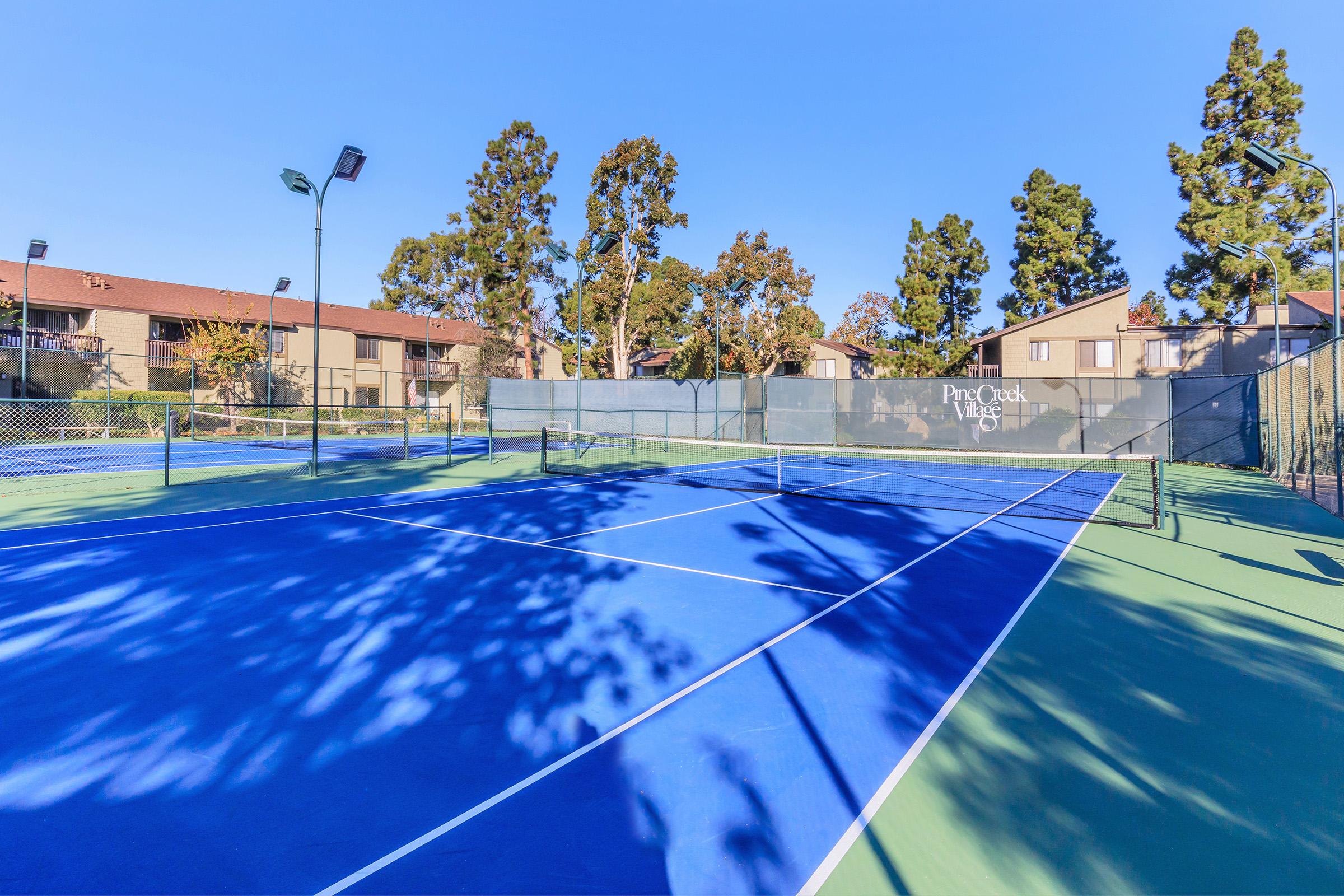 A well-maintained outdoor tennis court featuring blue and green surfaces, surrounded by trees and residential buildings in the background. The sky is clear and blue, with bright sunlight illuminating the area. Tennis court lights are visible, indicating potential for evening play.