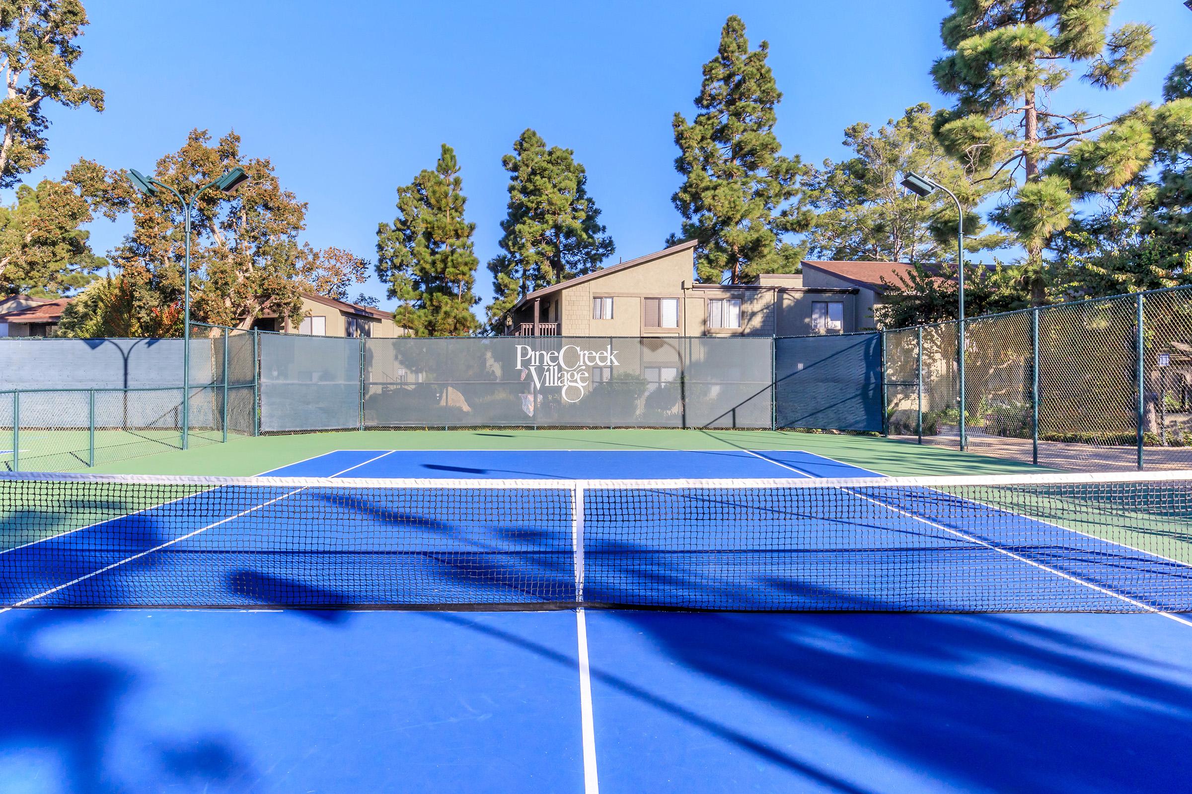 A well-maintained tennis court at Pine Creek Village, featuring a blue playing surface, surrounded by greenery and residential buildings. The clear blue sky adds to the vibrant atmosphere of the outdoor space, perfect for tennis games.