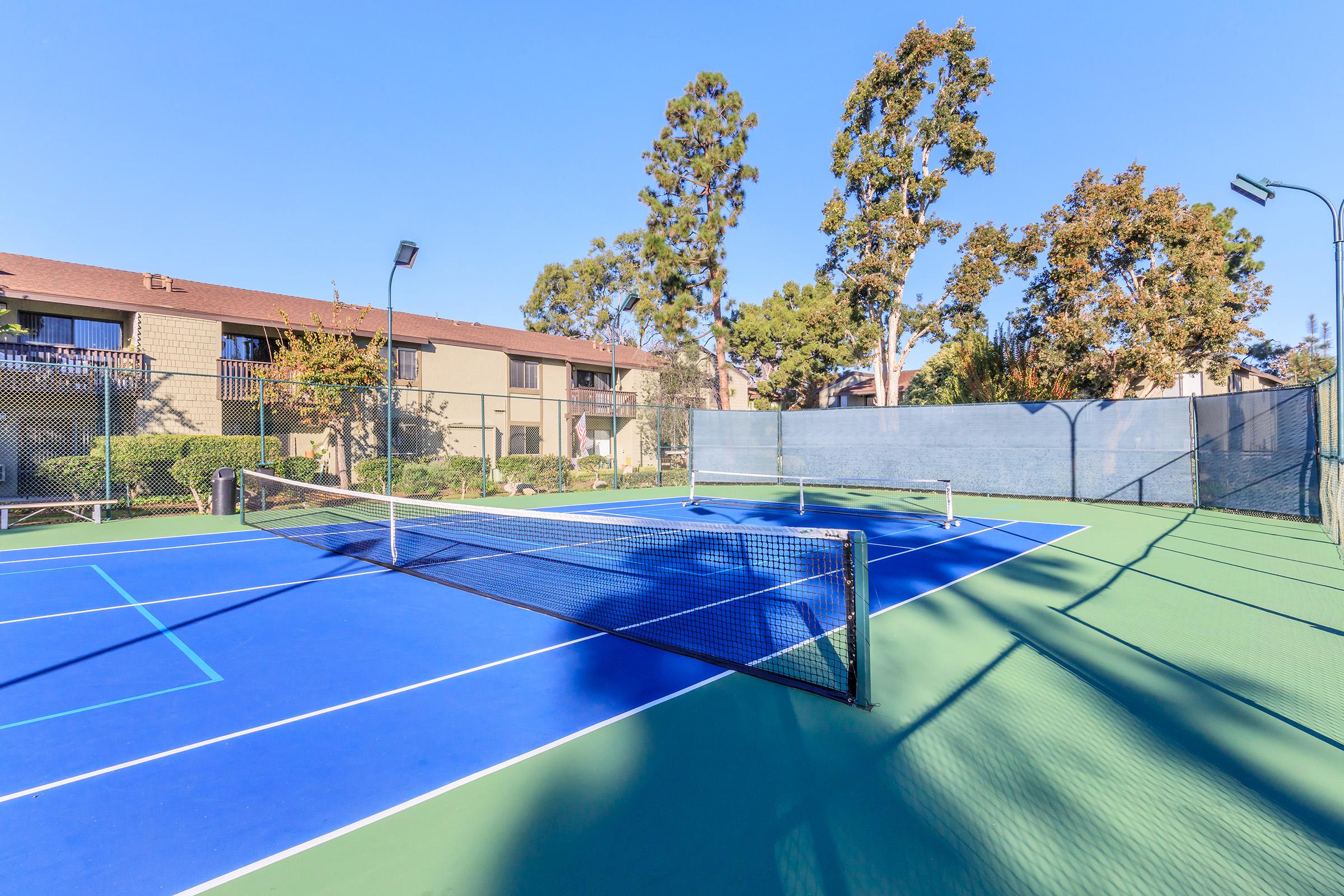 A bright tennis court featuring a blue surface with a net in the center, surrounded by green landscaping. The court is situated near apartment buildings under a clear blue sky, with tall trees casting shadows on the playing area.
