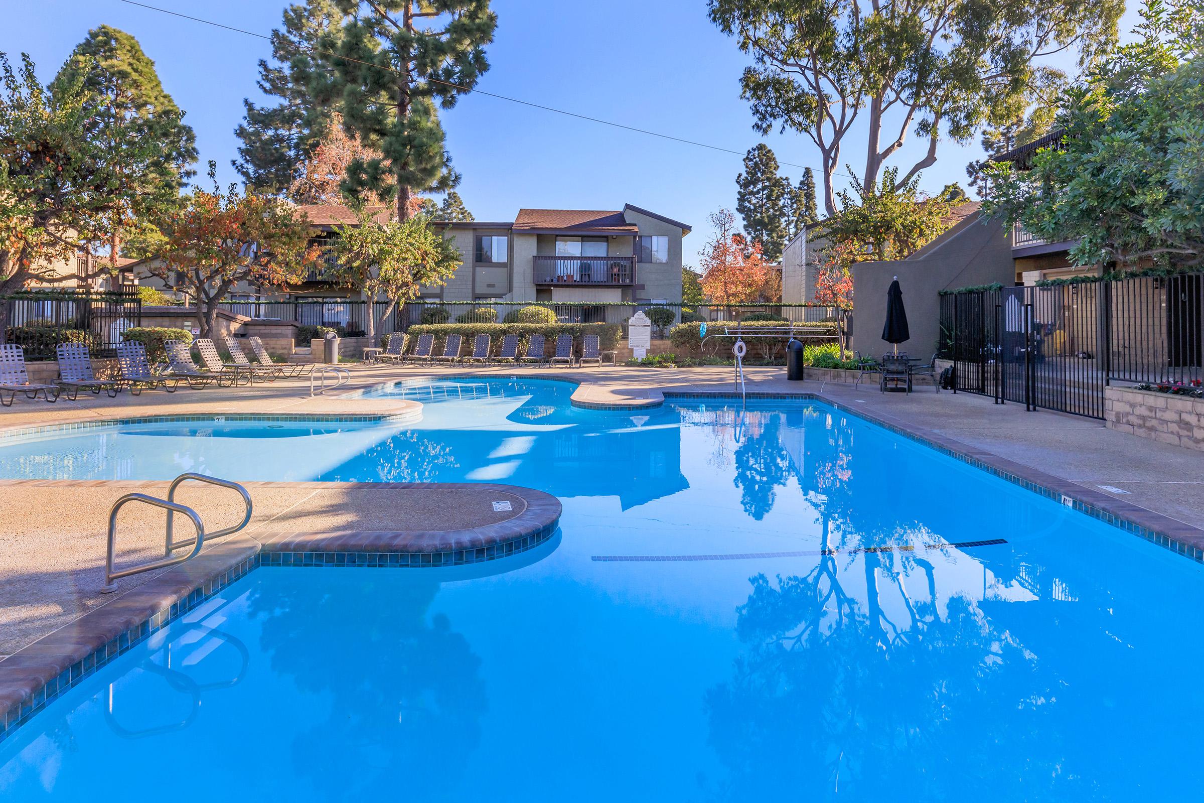 A clear blue swimming pool surrounded by landscaped greenery and trees. Lounge chairs line the pool area, with a few umbrellas scattered around. In the background, there are residential buildings. The scene is bright and inviting, reflecting a serene outdoor atmosphere.