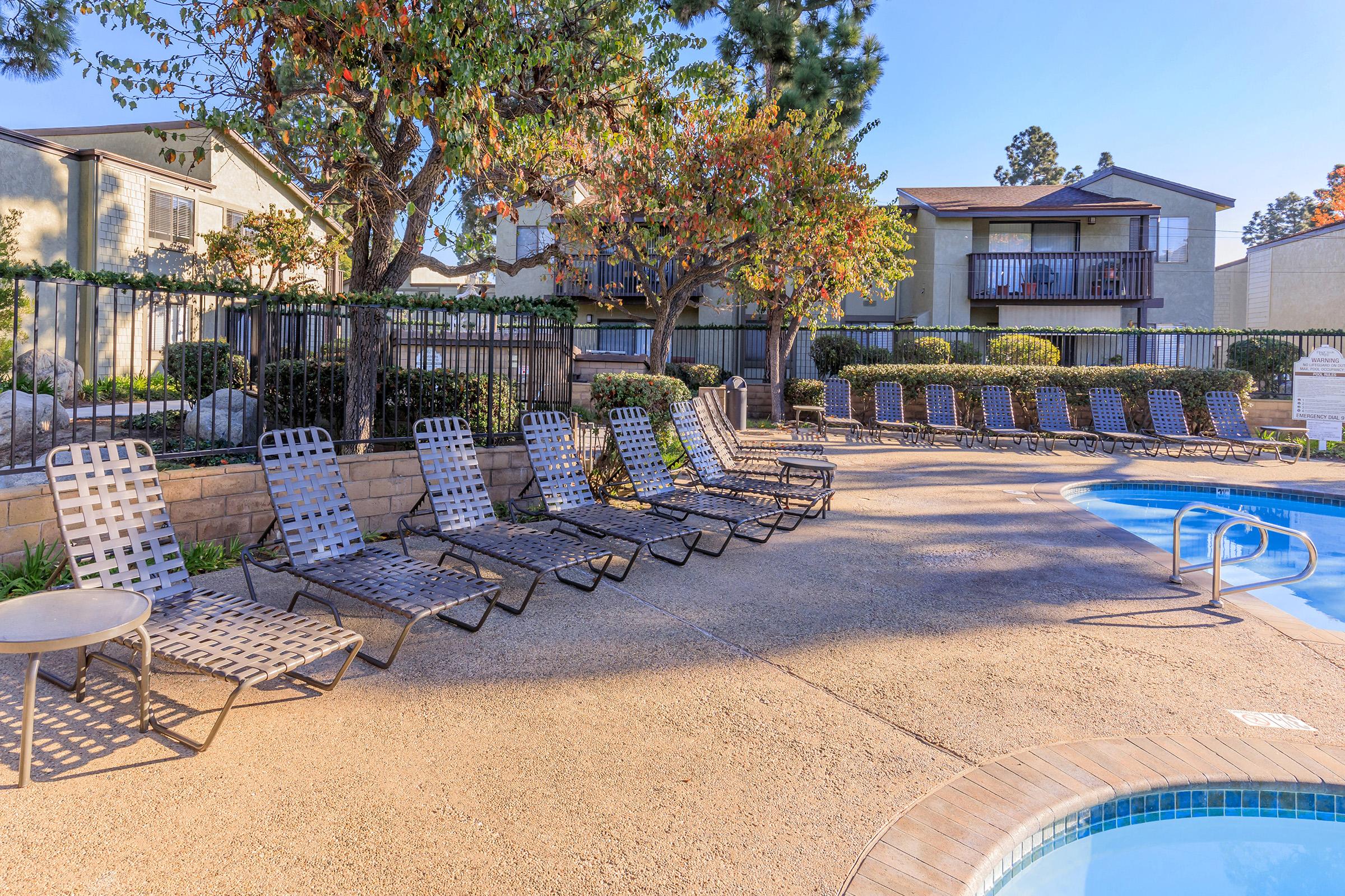 A sunny poolside scene featuring several lounge chairs arranged around a swimming pool, surrounded by trees and residential buildings. The area is neatly landscaped with shrubs and rocks, creating a relaxing atmosphere for leisure and relaxation.
