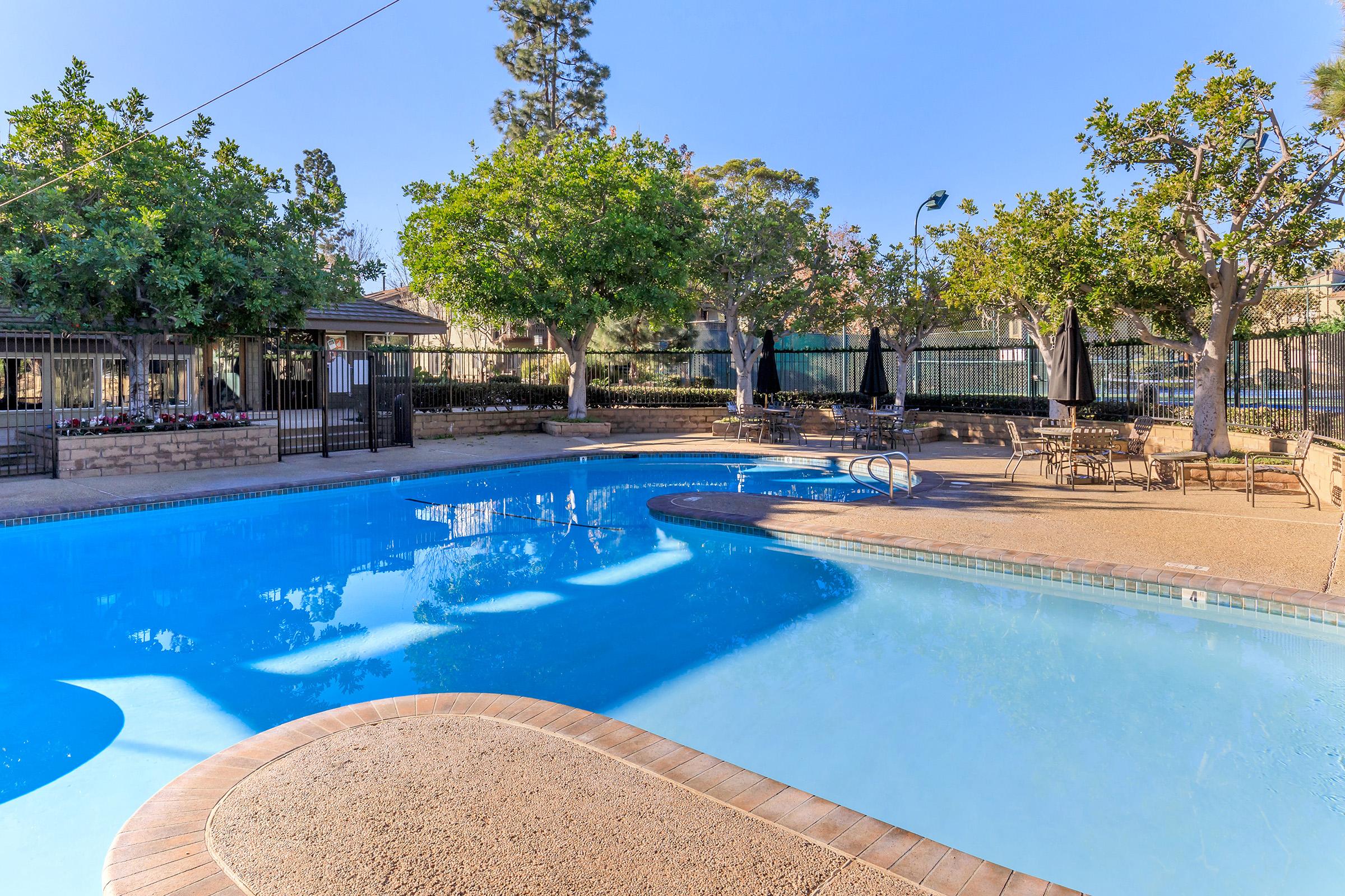 A spacious swimming pool surrounded by lush trees and lounge chairs. The pool area features a shallow section with steps leading in, and nearby are shaded seating areas with umbrellas. Bright blue skies and well-maintained landscaping create a welcoming atmosphere.