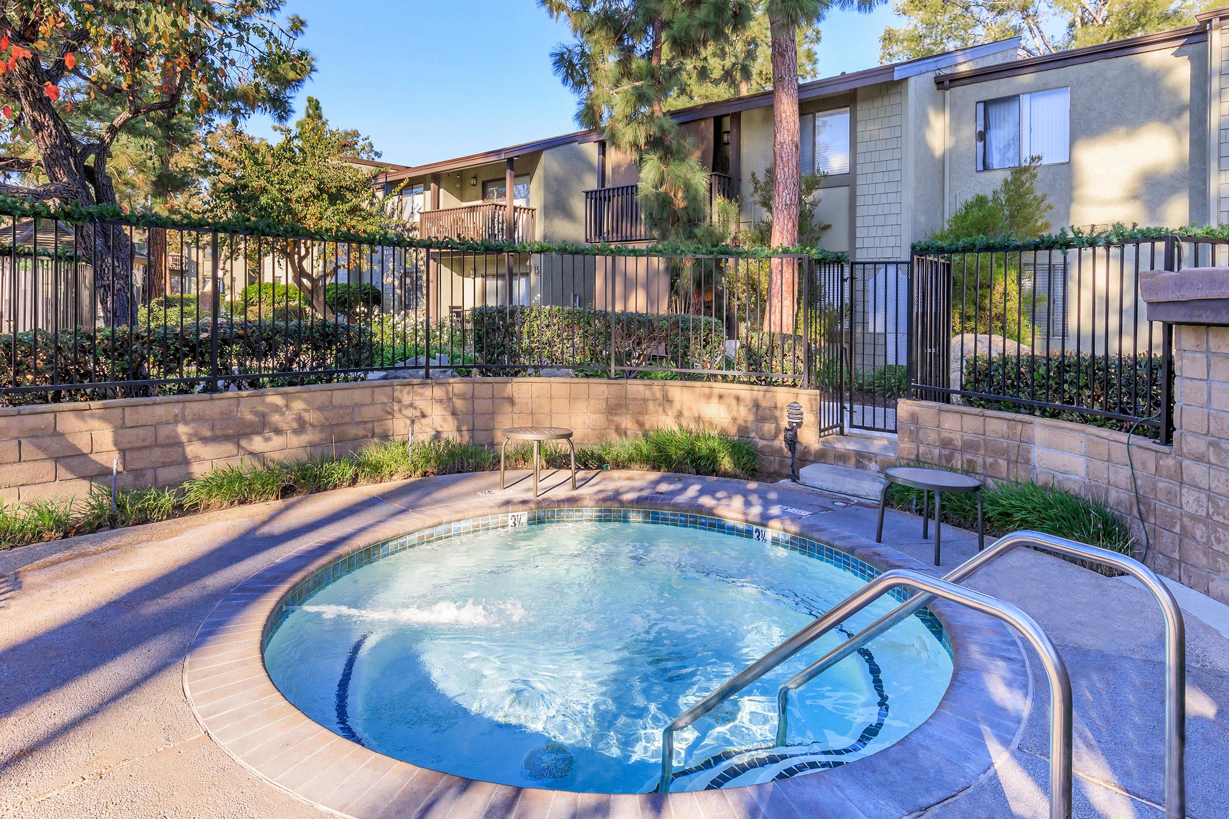A serene outdoor area featuring a circular hot tub, surrounded by greenery and a fence. In the background, there are well-maintained apartment buildings and trees, creating a peaceful residential environment. Sunlight illuminates the scene, enhancing the inviting atmosphere.