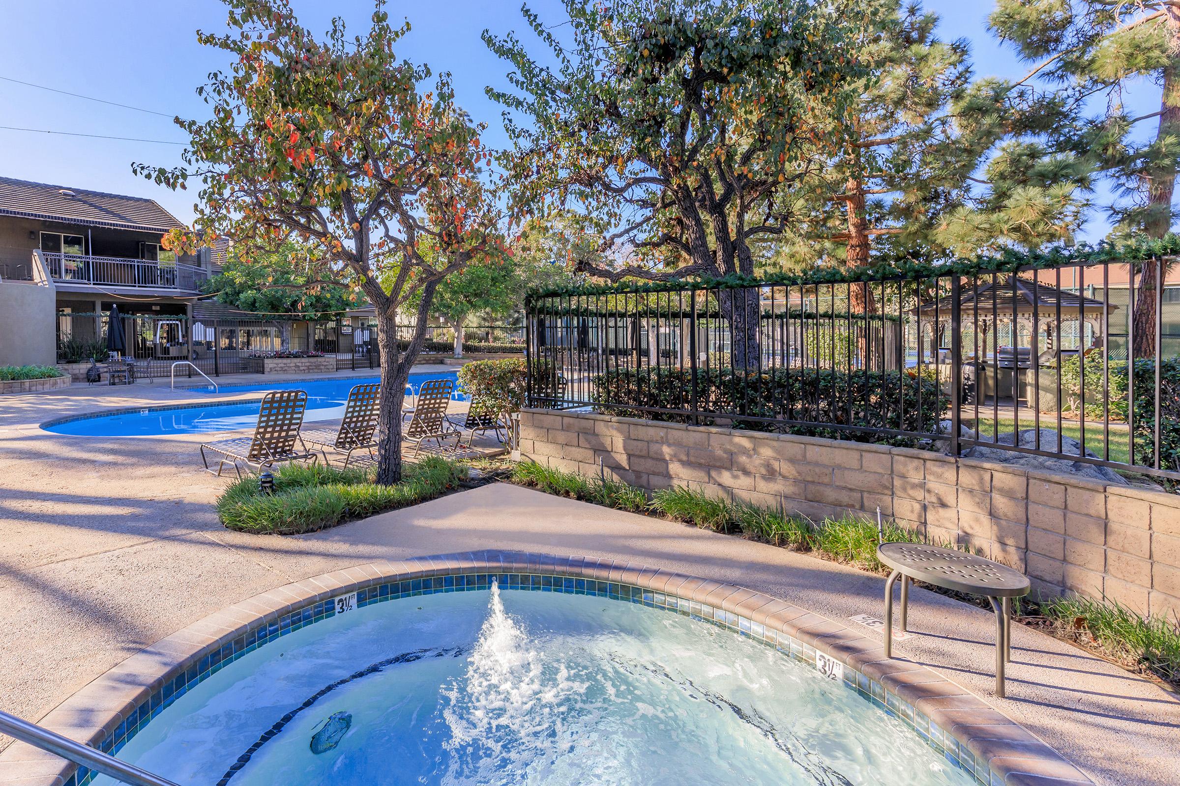 A serene outdoor area featuring a hot tub in the foreground, surrounded by greenery. In the background, there are two swimming pools, sun loungers, and well-maintained landscaping under clear blue skies. The scene conveys a relaxing atmosphere perfect for leisure and enjoyment.