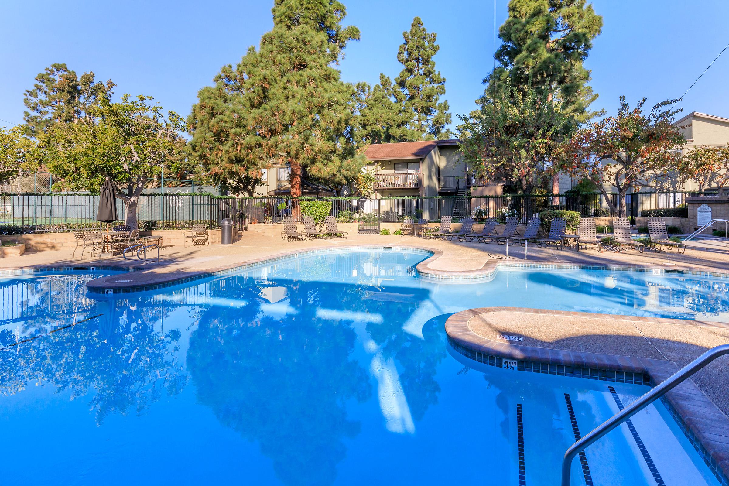 A sparkling blue swimming pool surrounded by lush greenery and trees. Lounge chairs are arranged around the pool area, with a few umbrellas providing shade. The sky is clear and sunny, creating a relaxing atmosphere in a residential setting.
