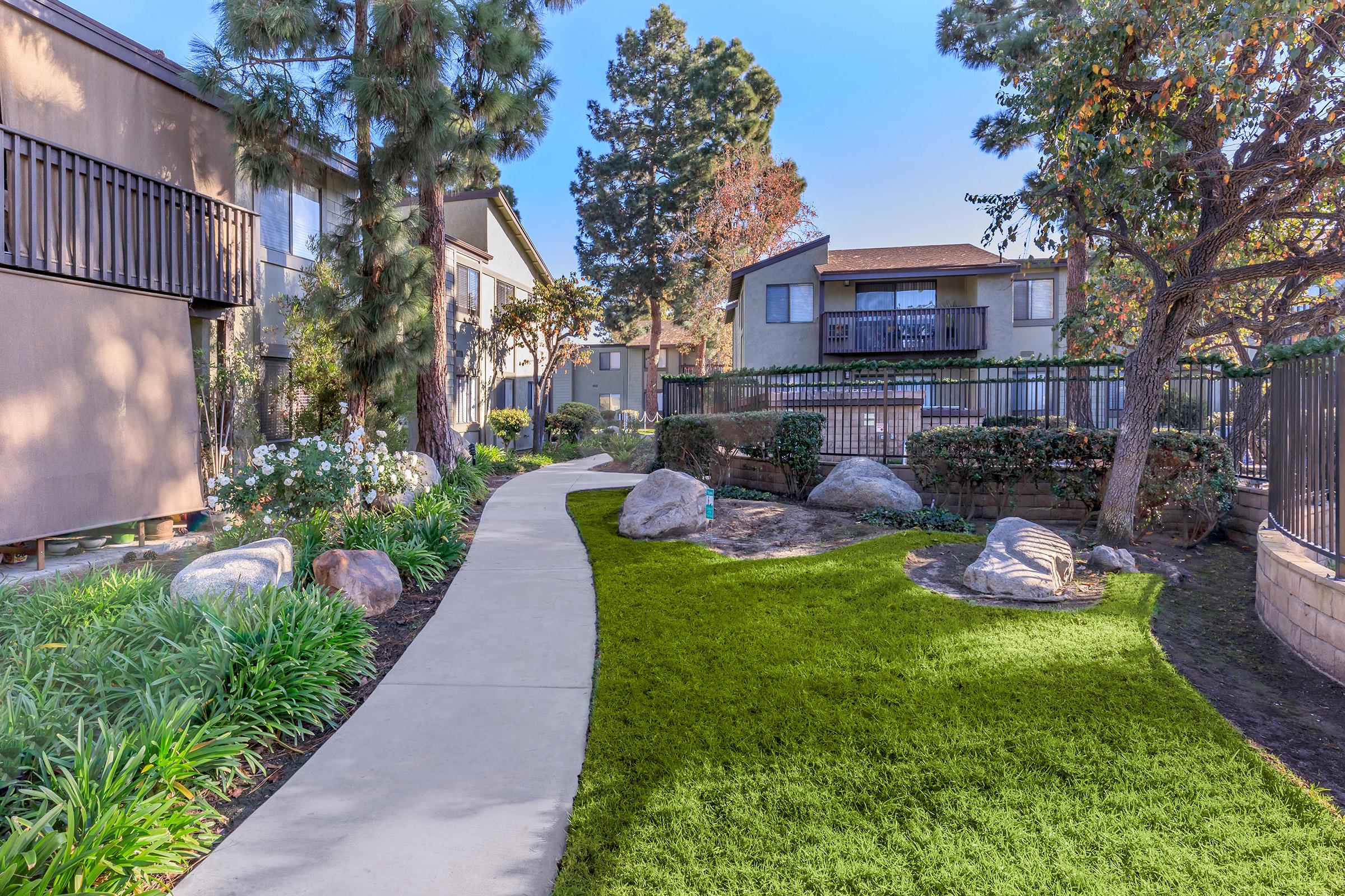 A landscaped pathway winds through a green lawn with vibrant shrubs and flowers. On either side, two multi-story residential buildings are visible, set among trees. The scene is bright and inviting, showcasing a well-maintained outdoor space.