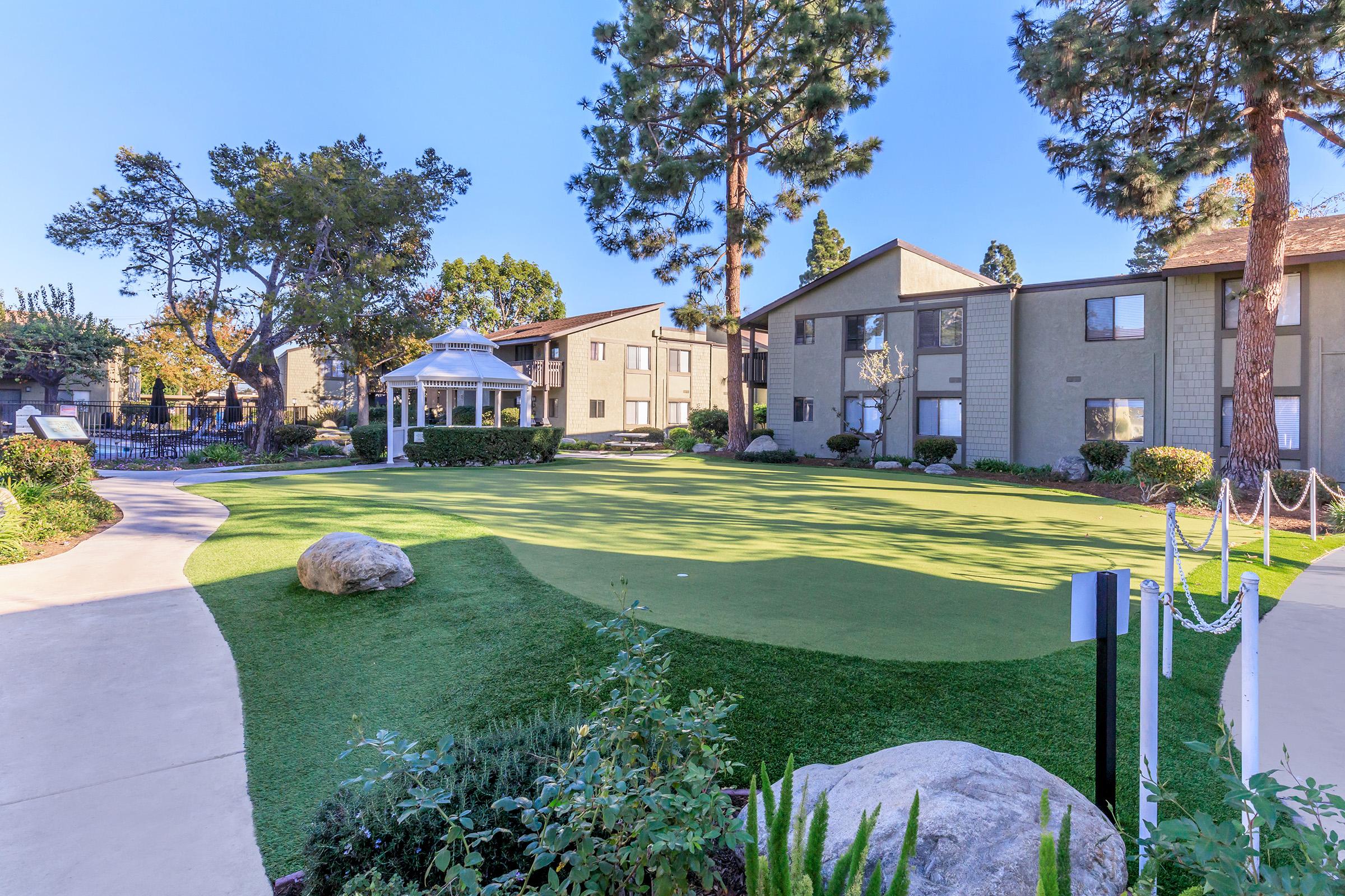 A well-maintained outdoor area featuring a putting green, surrounded by lush greenery and trees. In the background, there are residential buildings, and a gazebo is visible on the left side of the image, providing a relaxing space. The pathway is paved, adding to the inviting atmosphere.