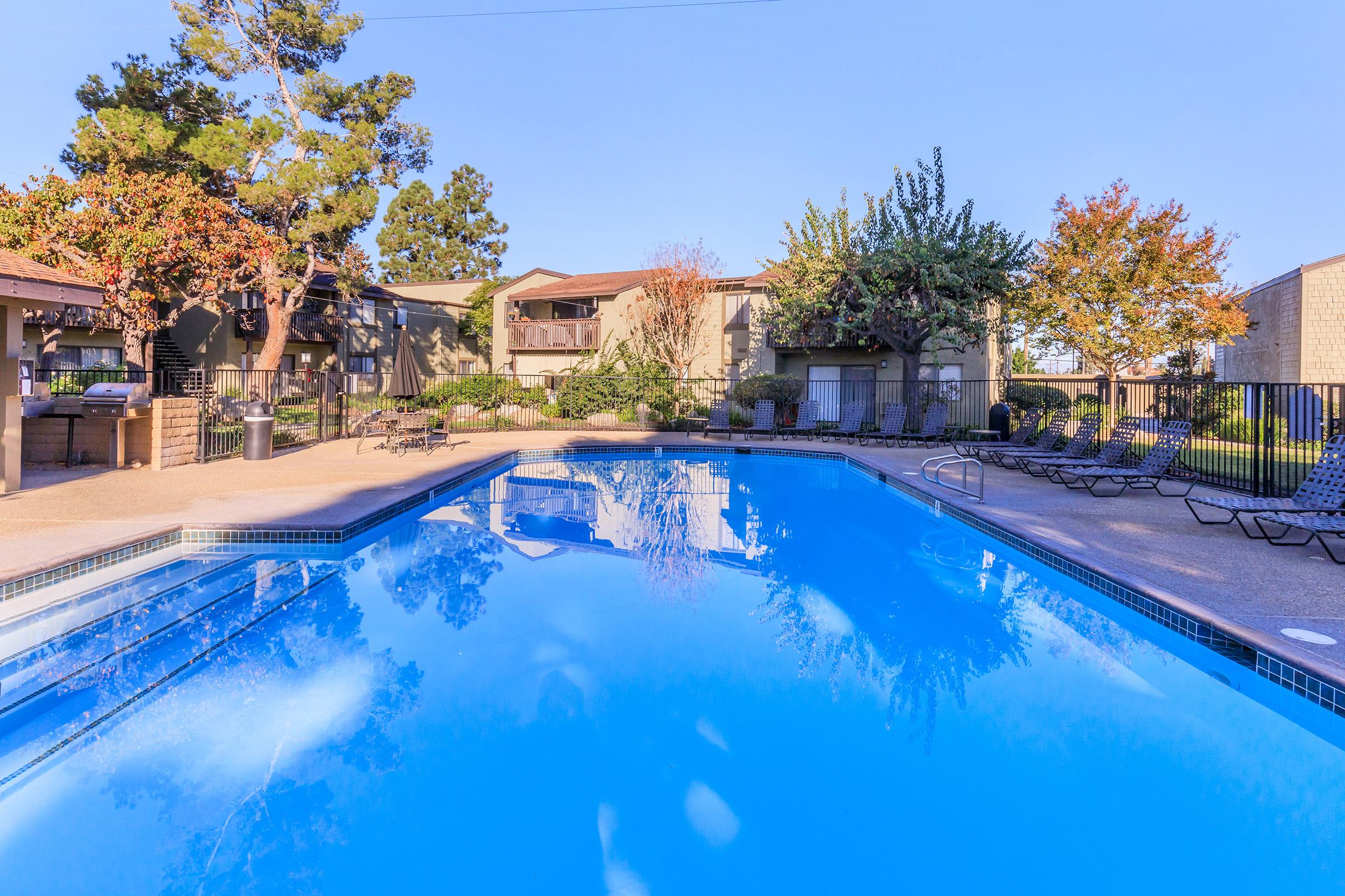 A clear blue swimming pool surrounded by lounge chairs and trees. In the background, there are two-story residential buildings with balconies and well-maintained landscaping, showcasing a peaceful outdoor setting. The sky is bright and sunny, adding to the inviting atmosphere.