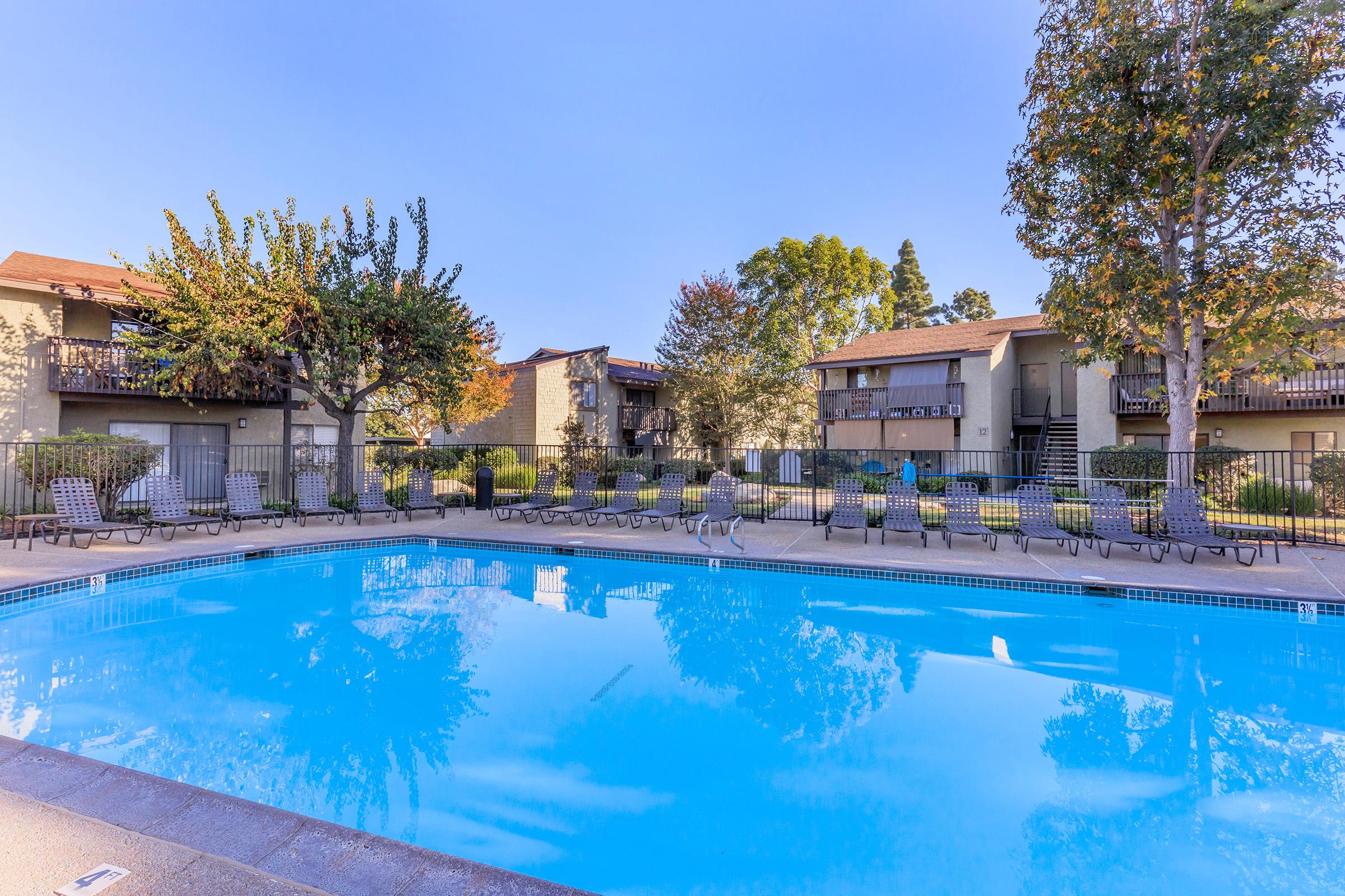 A clear blue swimming pool surrounded by lounge chairs, with two-story apartment buildings in the background. Lush trees and shrubs frame the scene, reflecting a bright, sunny day. The setting conveys a relaxing outdoor atmosphere typical of a residential community.