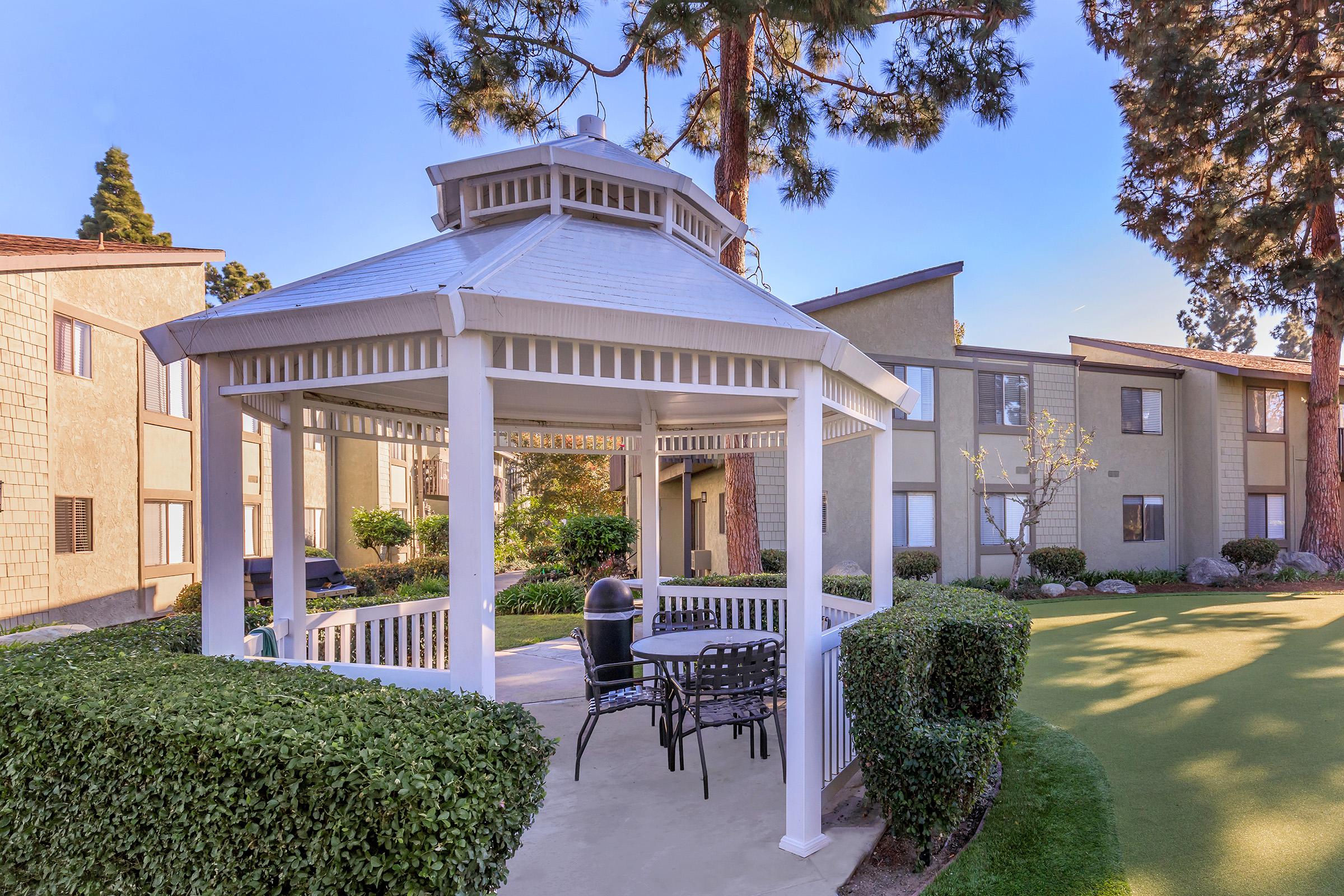 A peaceful outdoor gazebo surrounded by neatly trimmed hedges and a manicured lawn. In the gazebo, there are black chairs and a table, providing a cozy place to relax. Behind it, there are apartment buildings and tall pine trees under a clear blue sky, creating a serene residential atmosphere.