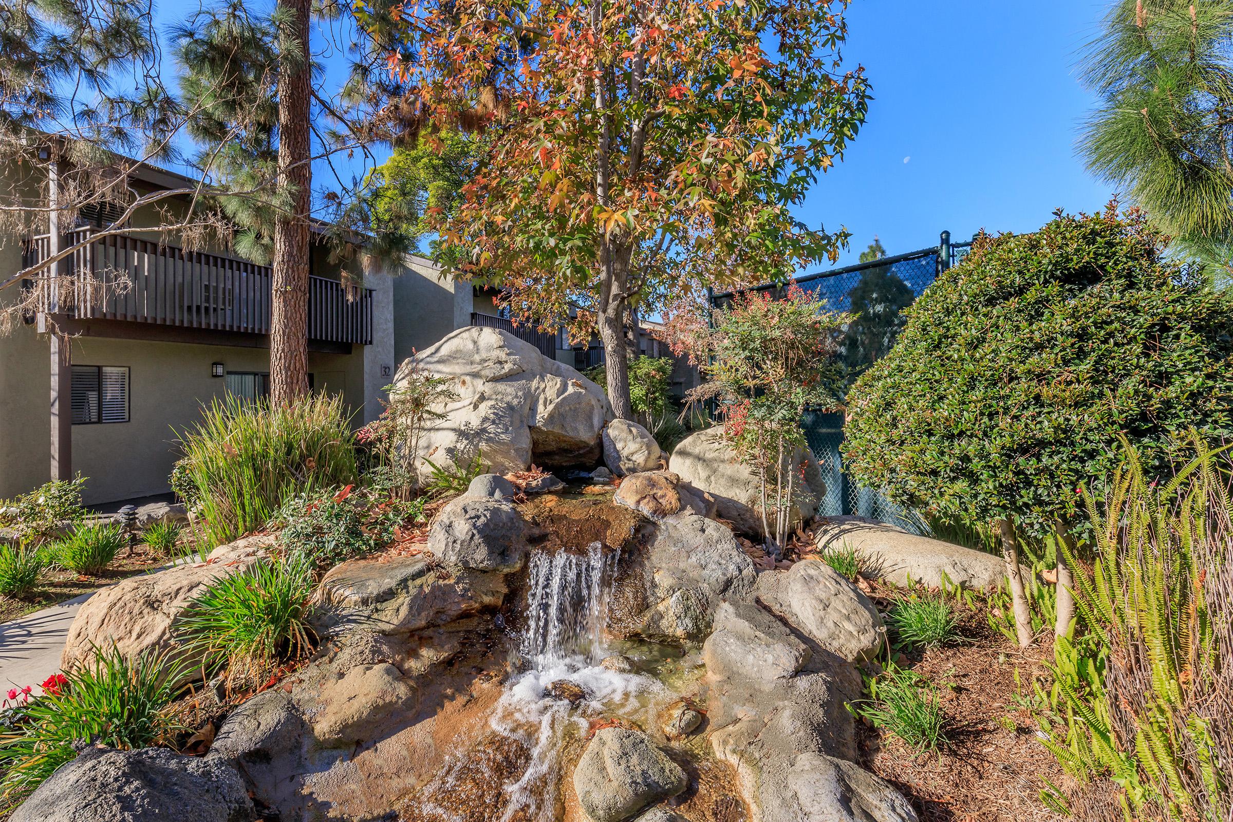 A serene outdoor scene featuring a small waterfall flowing over smooth stones, surrounded by lush green plants and a tree with autumn foliage. In the background, there are residential buildings and a blue sky, contributing to a peaceful garden atmosphere.