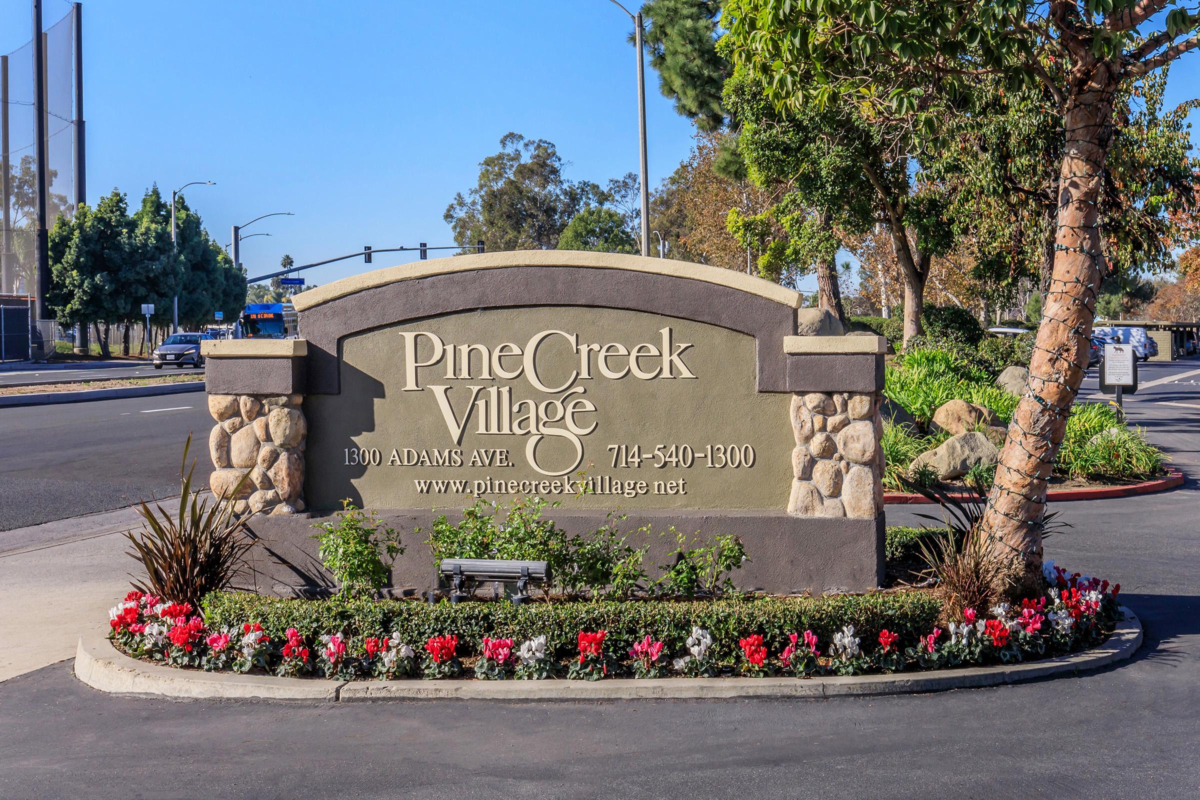 Sign for PineCreek Village located at 1300 Adams Ave, featuring contact number 714-540-1300. The sign is surrounded by colorful flowers and greenery, set against a clear sky.