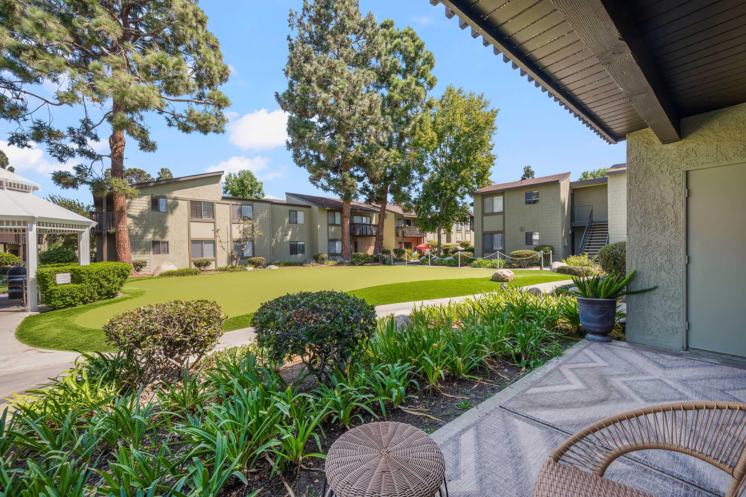 A landscaped courtyard featuring manicured grass and colorful plants, surrounded by two-story apartment buildings. The scene includes palm trees and garden furniture, under a clear blue sky, creating a serene and inviting outdoor space.
