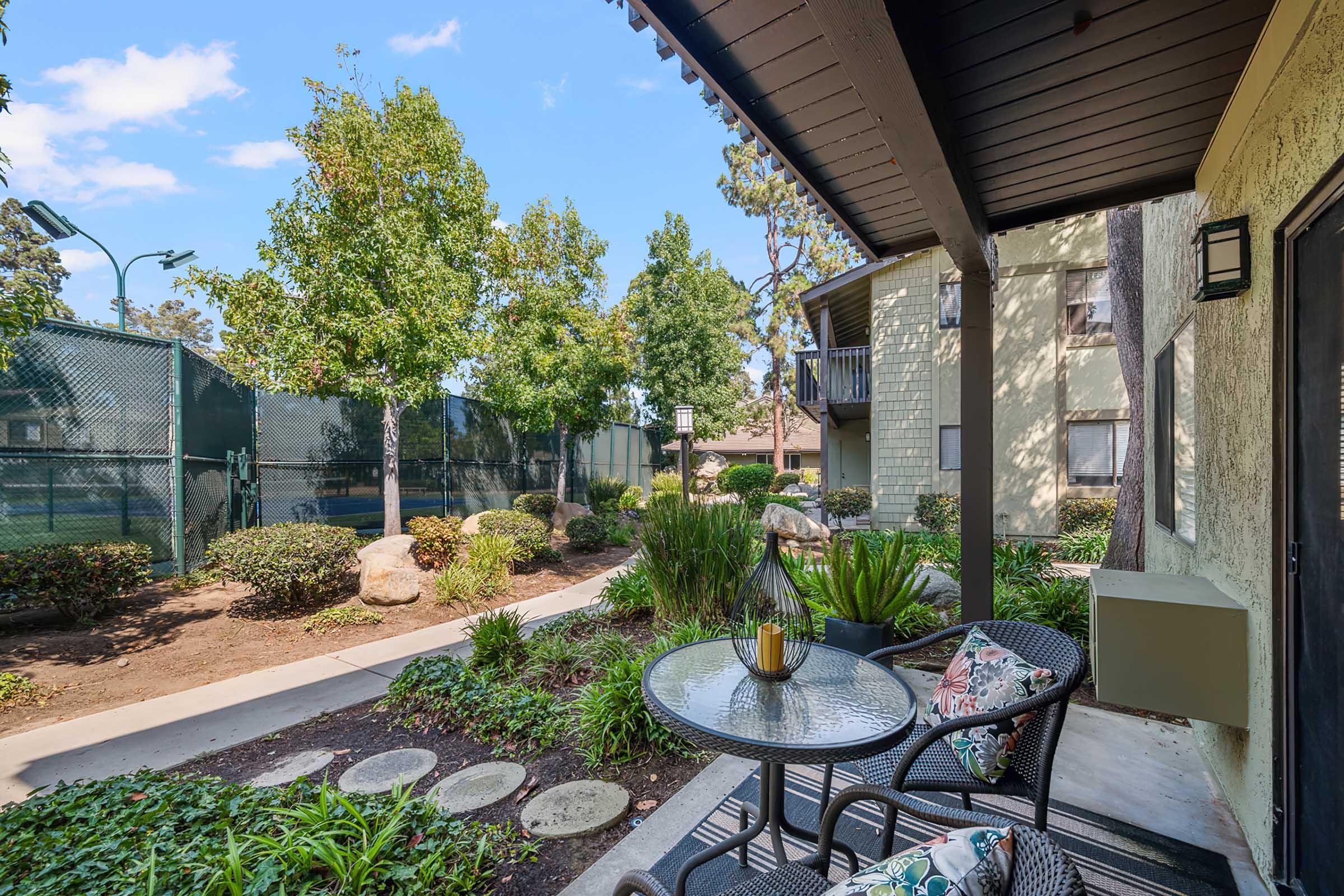 A cozy outdoor patio features a small round table with decorative cushions and a glass of drink, surrounded by lush greenery and colorful plants. In the background, a well-maintained pathway leads to a tennis court, with tall trees providing shade and a peaceful atmosphere.