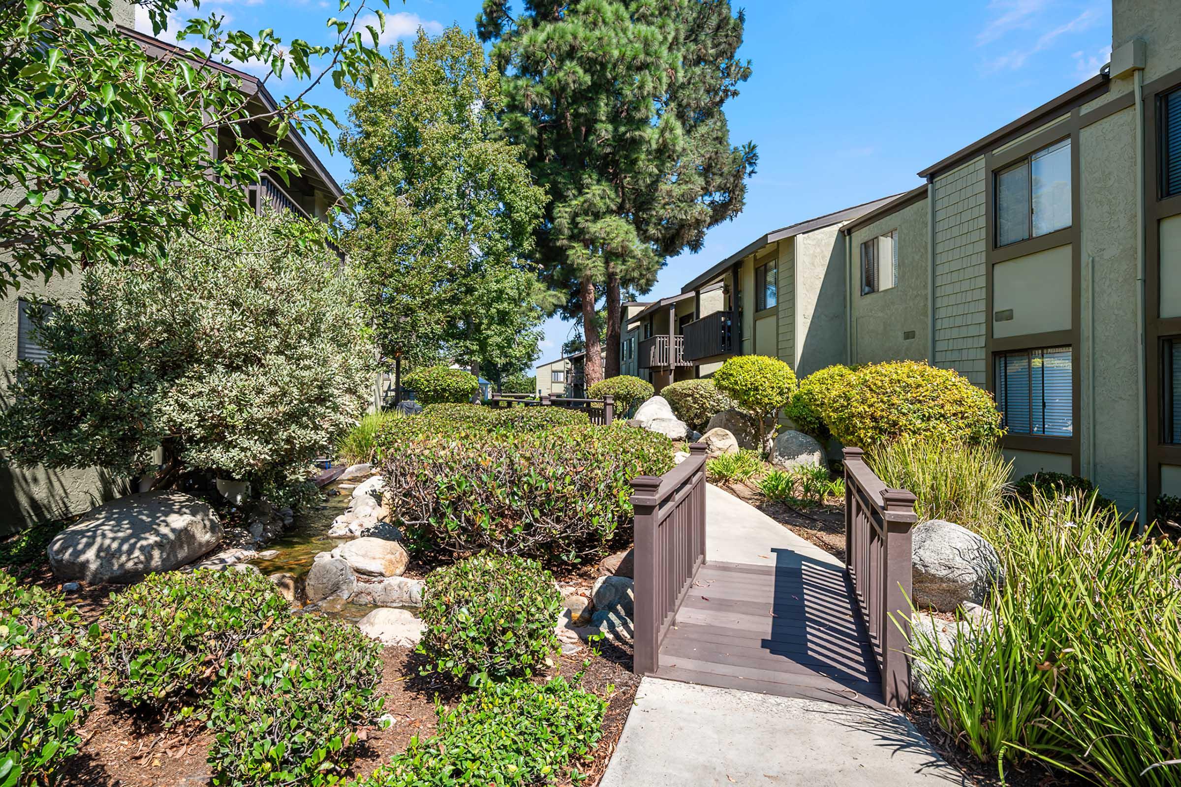 A peaceful pathway through a landscaped area featuring shrubs, rocks, and trees, leading to residential buildings. The scene is sunny and inviting, with well-maintained greenery and a wooden bridge over a small water feature.