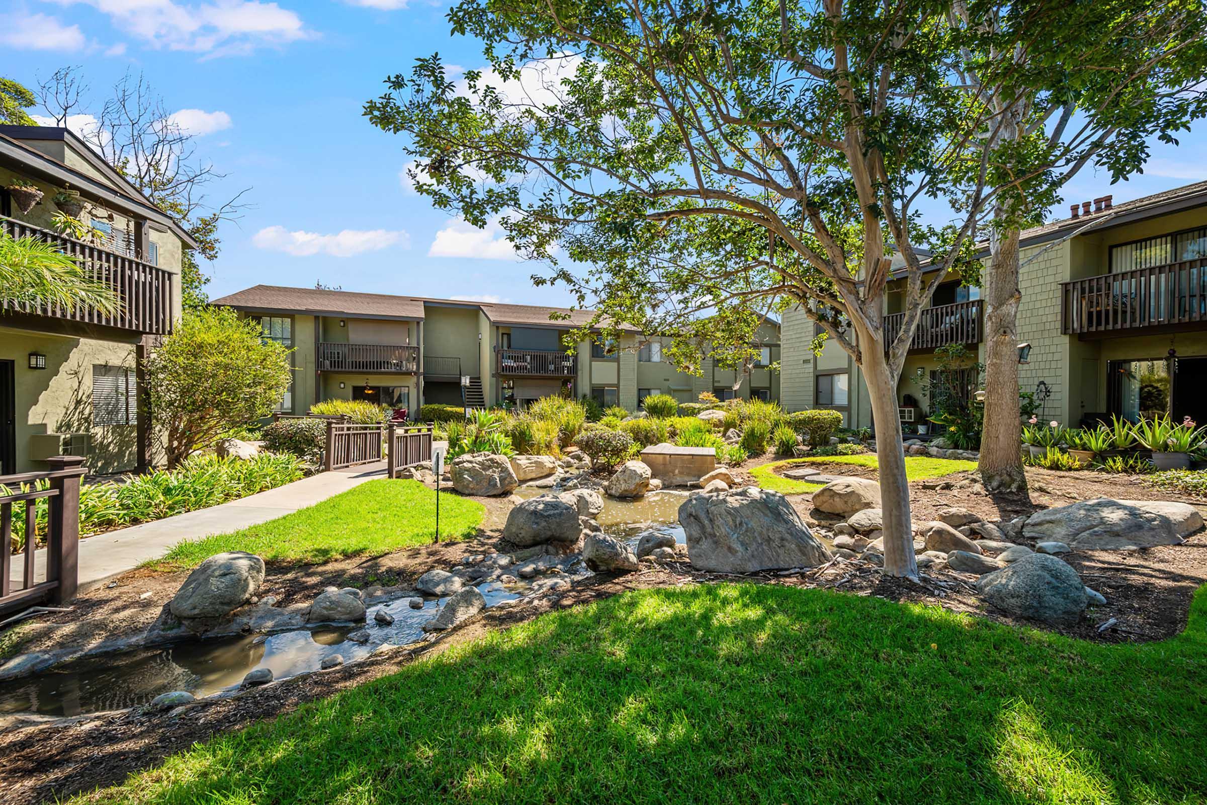 A landscaped courtyard of an apartment complex featuring green grass, large rocks, a small water feature, and surrounding trees. The buildings are two stories high with balconies, providing a serene environment for residents. Clear blue sky adds to the inviting atmosphere.