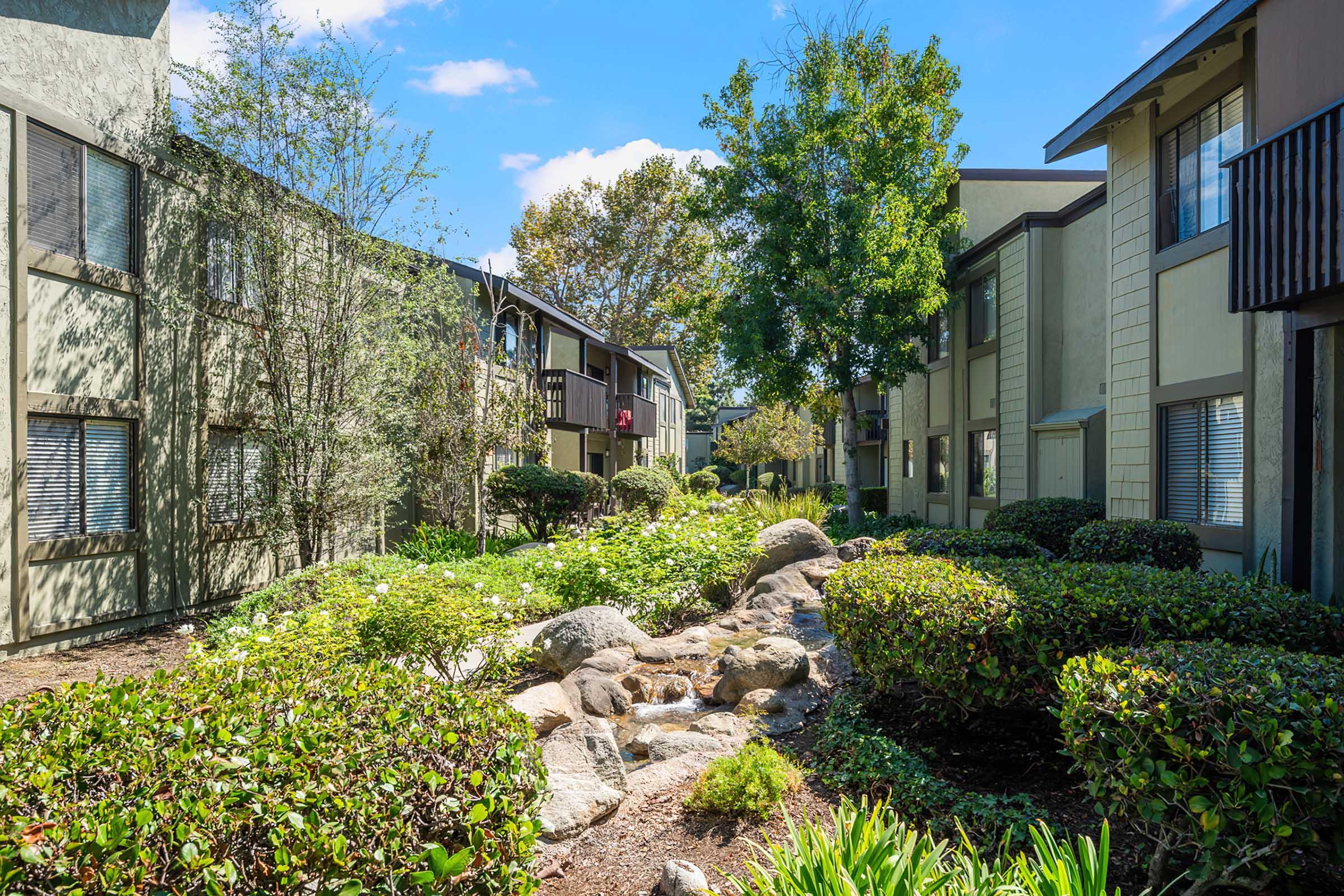 A landscaped pathway lined with greenery and flowering plants, leading between two apartment buildings. The scene features rocks, shrubs, and trees under a clear blue sky, creating a tranquil residential setting.