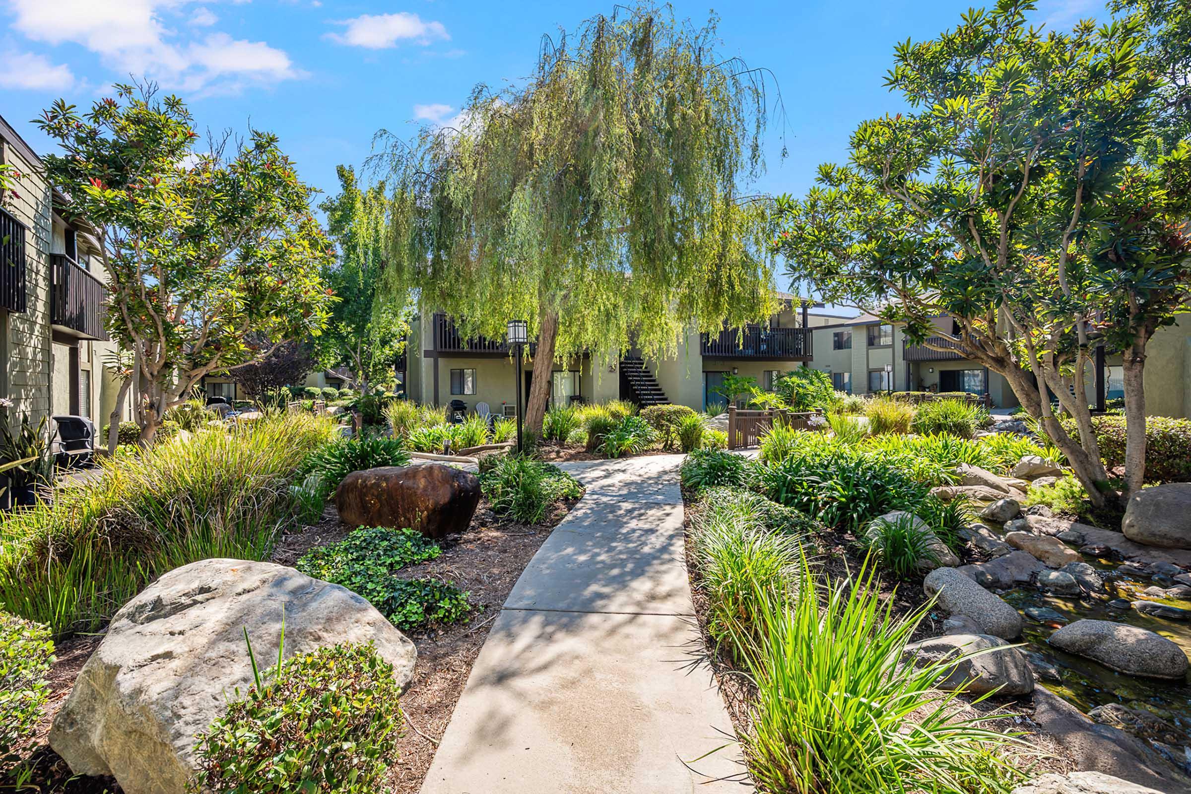 A landscaped garden pathway meanders through lush greenery, dotted with rocks and a small pond. Surrounding buildings are partially visible in the background, under a clear blue sky. The scene evokes a tranquil outdoor space, ideal for relaxation and nature appreciation.