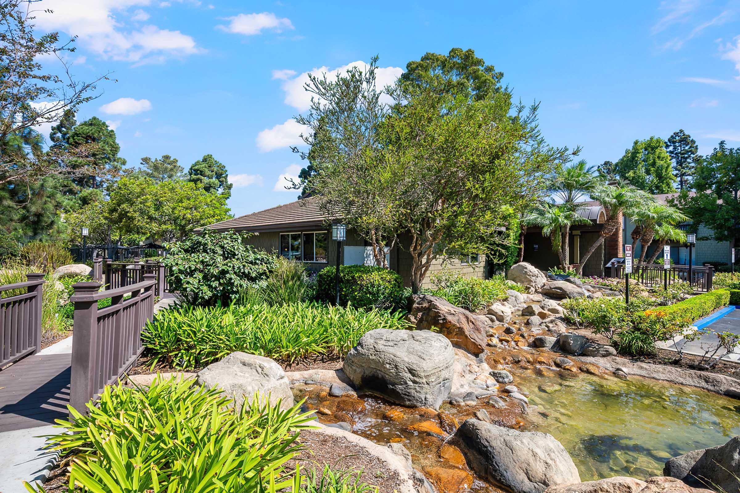 A tranquil landscape featuring a small rock-lined pond surrounded by lush greenery, including bushes and trees. A wooden bridge crosses over the pond, leading to a building in the background. The setting is bright and sunny, with a clear blue sky.
