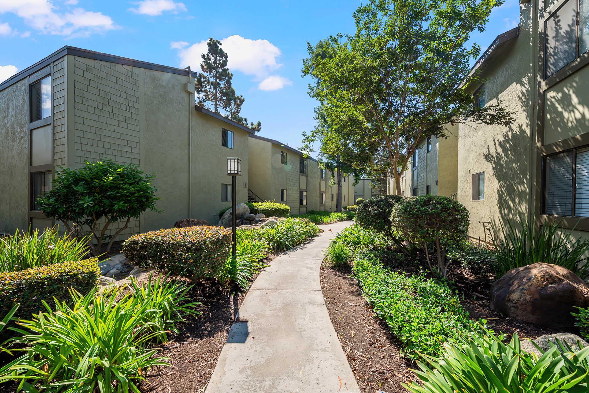 A well-maintained pathway meanders through a landscaped area surrounded by two-story apartment buildings. Lush greenery, including shrubs and flower beds, adorns the sides of the path, creating a serene and inviting atmosphere under a bright blue sky.