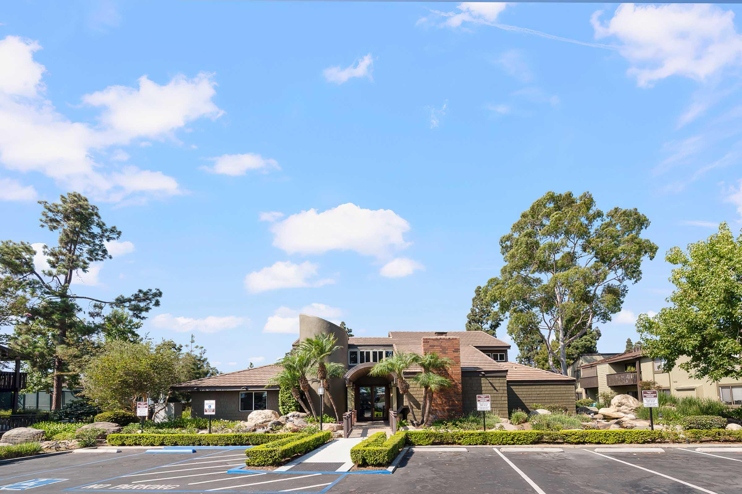 A modern building with a combination of stone and wood architecture, surrounded by well-maintained landscaping and palm trees. The sky is clear with fluffy white clouds. A parking lot in front features marked spaces, indicating a welcoming entrance to the facility.