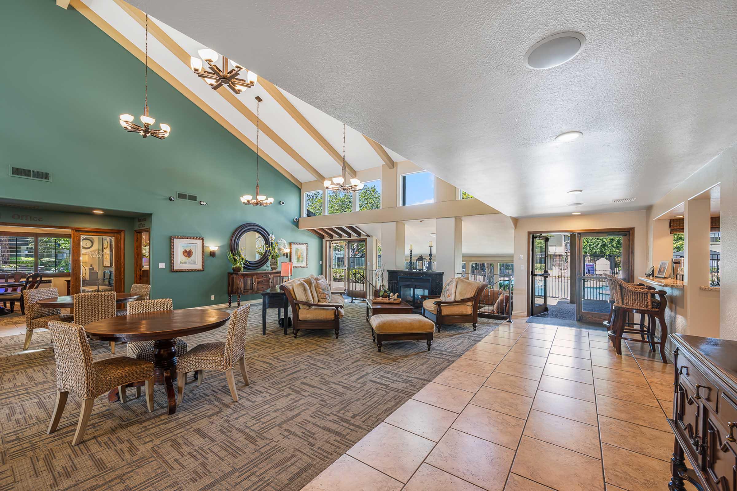 A spacious, well-lit lobby featuring a mix of modern and comfortable seating arrangements, including woven chairs and a wooden table. Large windows provide natural light, and decorative chandeliers hang from a high ceiling. The area has a welcoming atmosphere with stylish decor and access to an outdoor space.