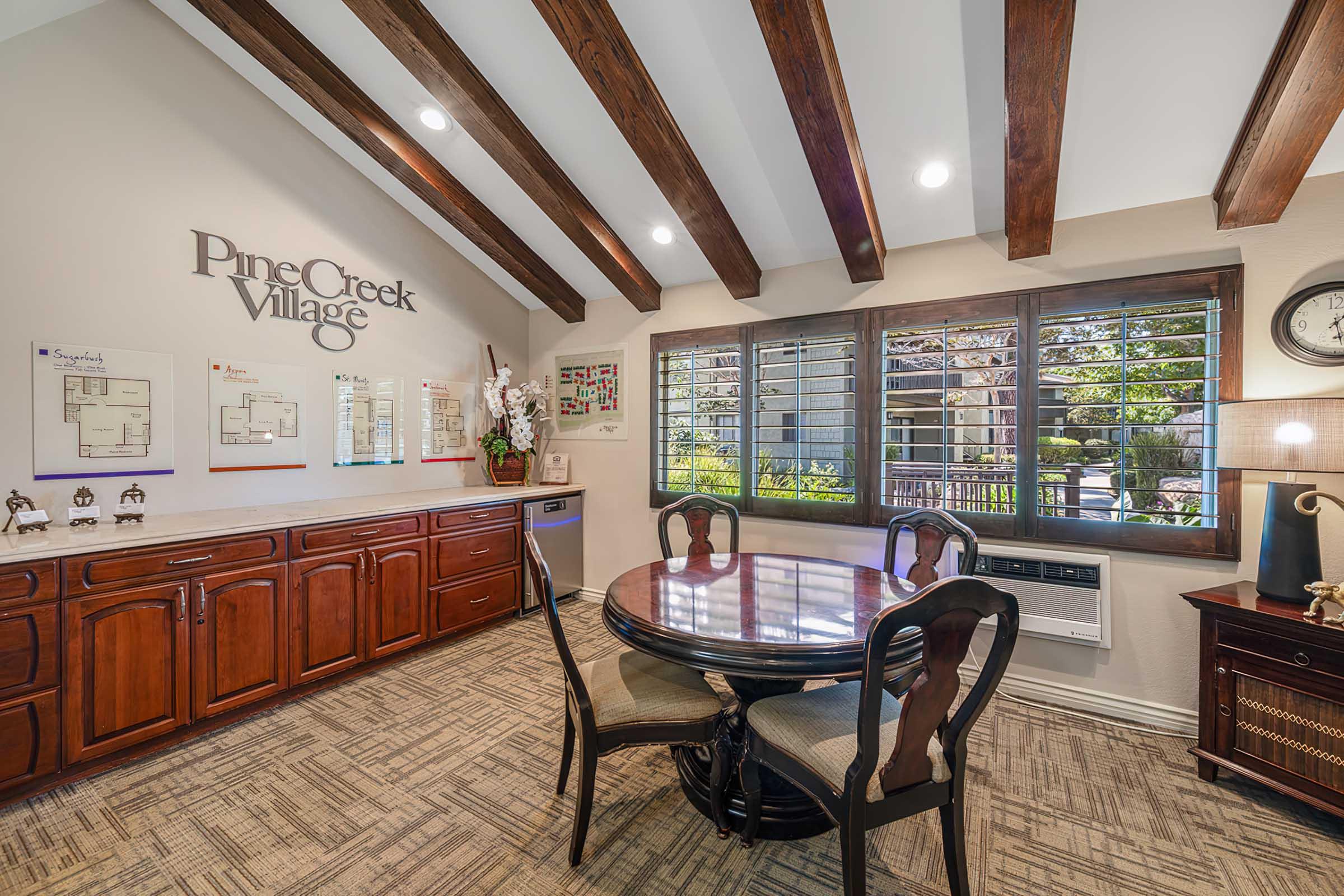 A cozy interior of a room featuring wooden beams on the ceiling, a round dining table with upholstered chairs, and dark wood cabinets. The wall is adorned with a sign reading "Pine Creek Village" and a few framed documents. Large windows allow natural light, showcasing a view of greenery outside.