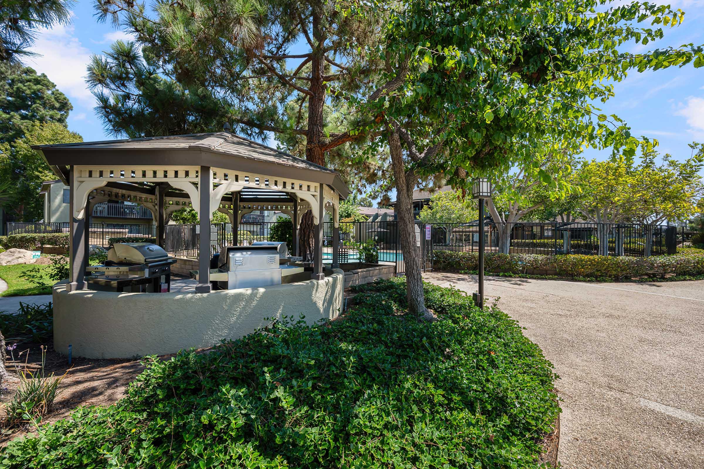A gazebo with a grill surrounded by greenery, situated in a landscaped area. In the background, a fence and swimming pool are visible. The scene is sunny with blue skies and trees providing shade. The walkway is made of gravel and features well-maintained plants.