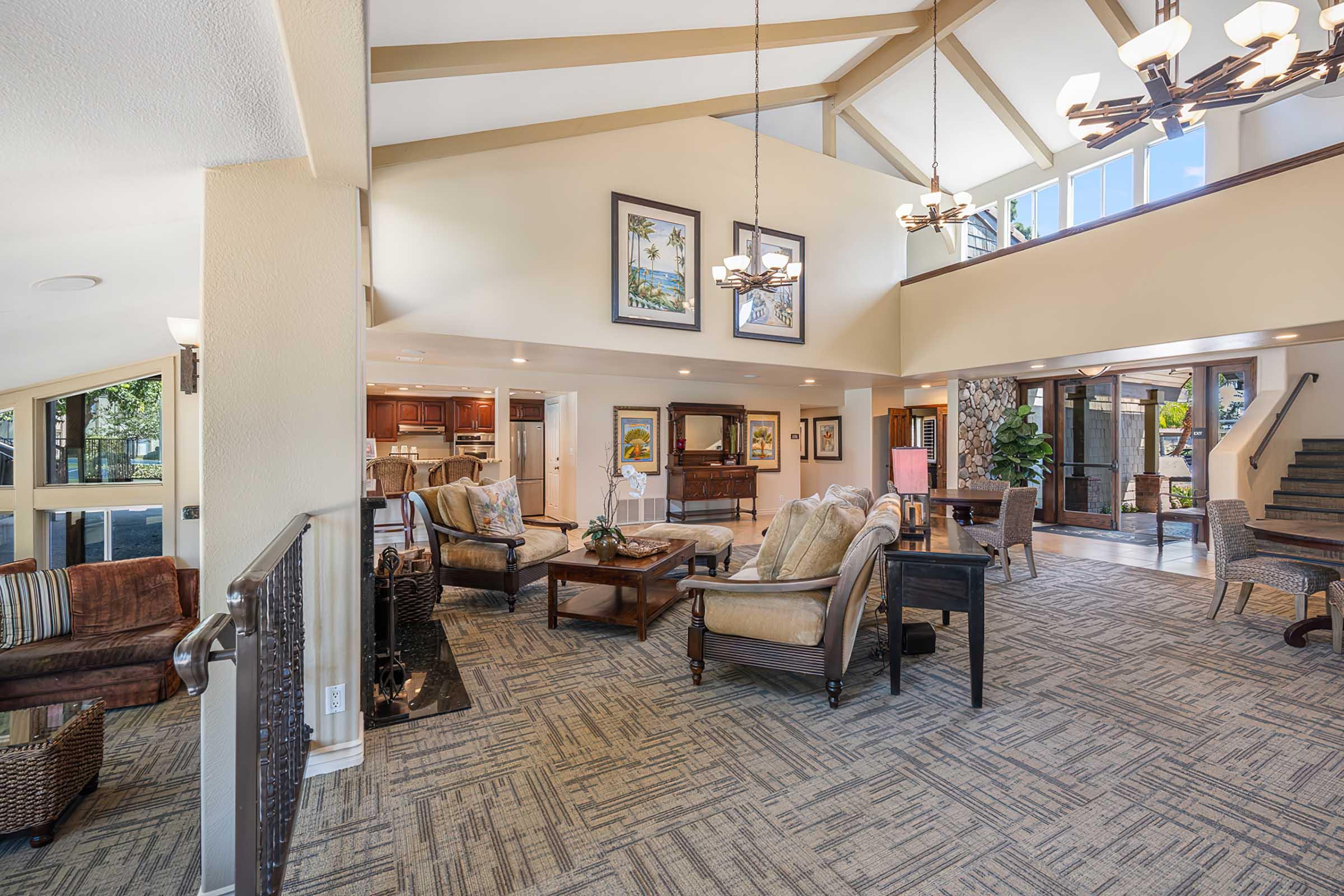 Spacious and modern living area featuring high vaulted ceilings, large windows, and a cozy seating arrangement. The decor includes elegant furniture, artworks on the walls, and a blend of warm and neutral colors. A staircase leads to an upper level, with an open layout connecting to a dining and kitchen area.