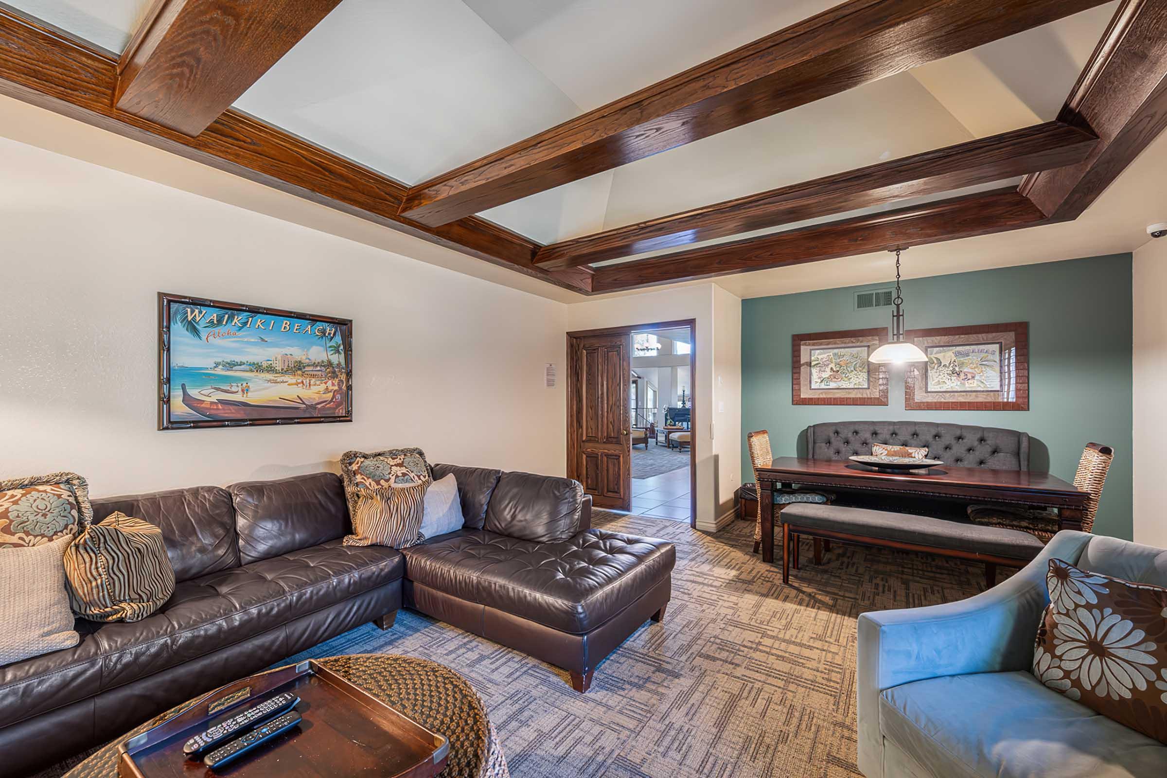 A cozy living area featuring a dark leather sectional sofa with decorative pillows, a round coffee table, and a light blue accent chair. There is a dining space with a dark wooden table and upholstered chairs, complemented by a wall painted in a calming green shade. Wooden beams add architectural interest to the ceiling.