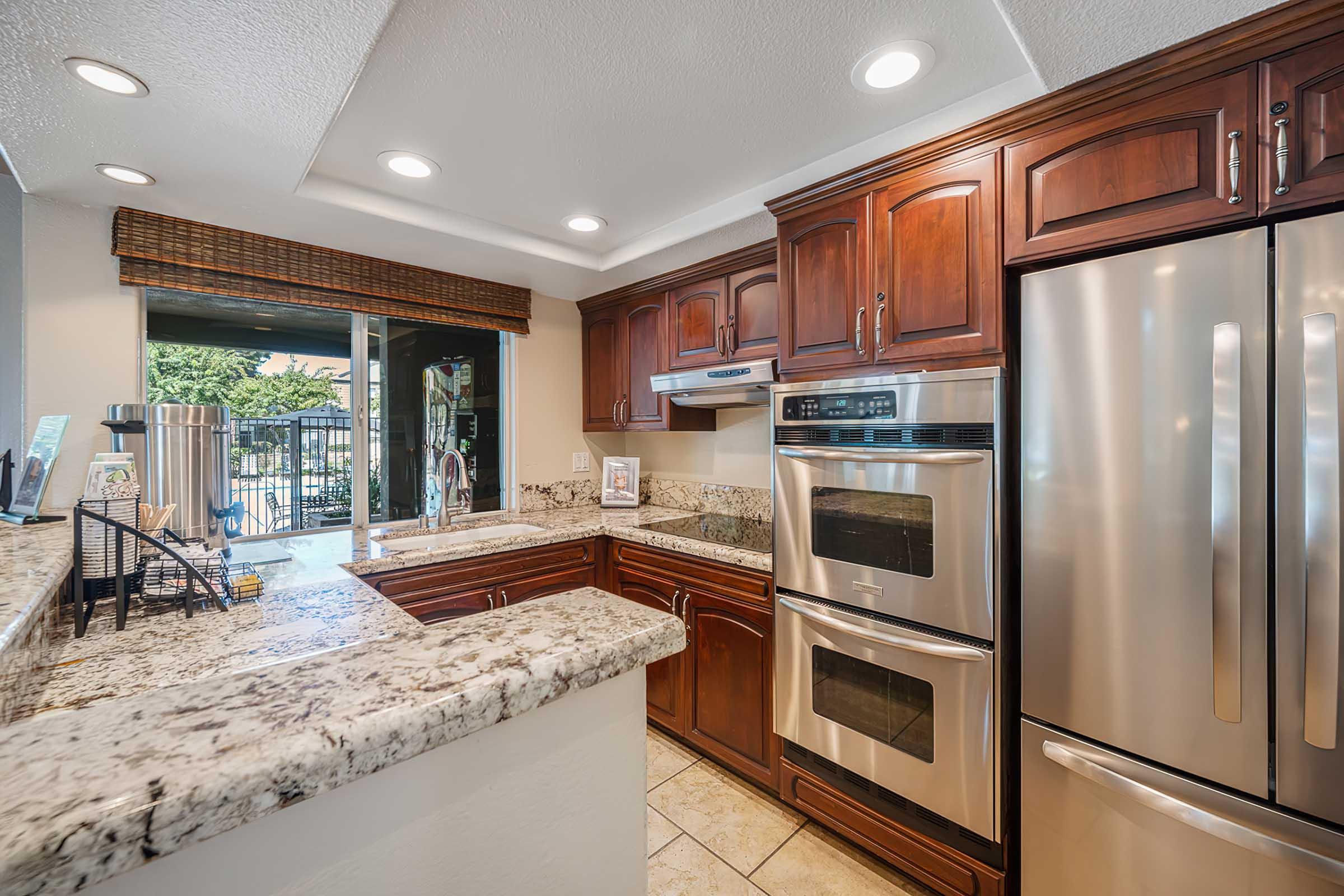 A modern kitchen featuring dark wood cabinets, stainless steel appliances, double ovens, and a large granite countertop. Natural light streams in through a window, highlighting the elegant design. The space includes a view of an outdoor area, enhancing the inviting atmosphere.