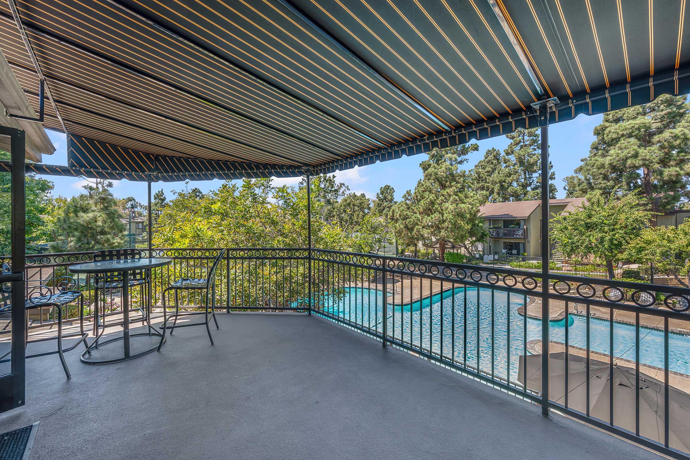 A spacious balcony with a wrought iron railing offers a view of a swimming pool surrounded by greenery. The awning above provides shade, and there are two black chairs and a small table set on the balcony, creating a relaxing outdoor space. The scene captures a sunny day in a residential area.