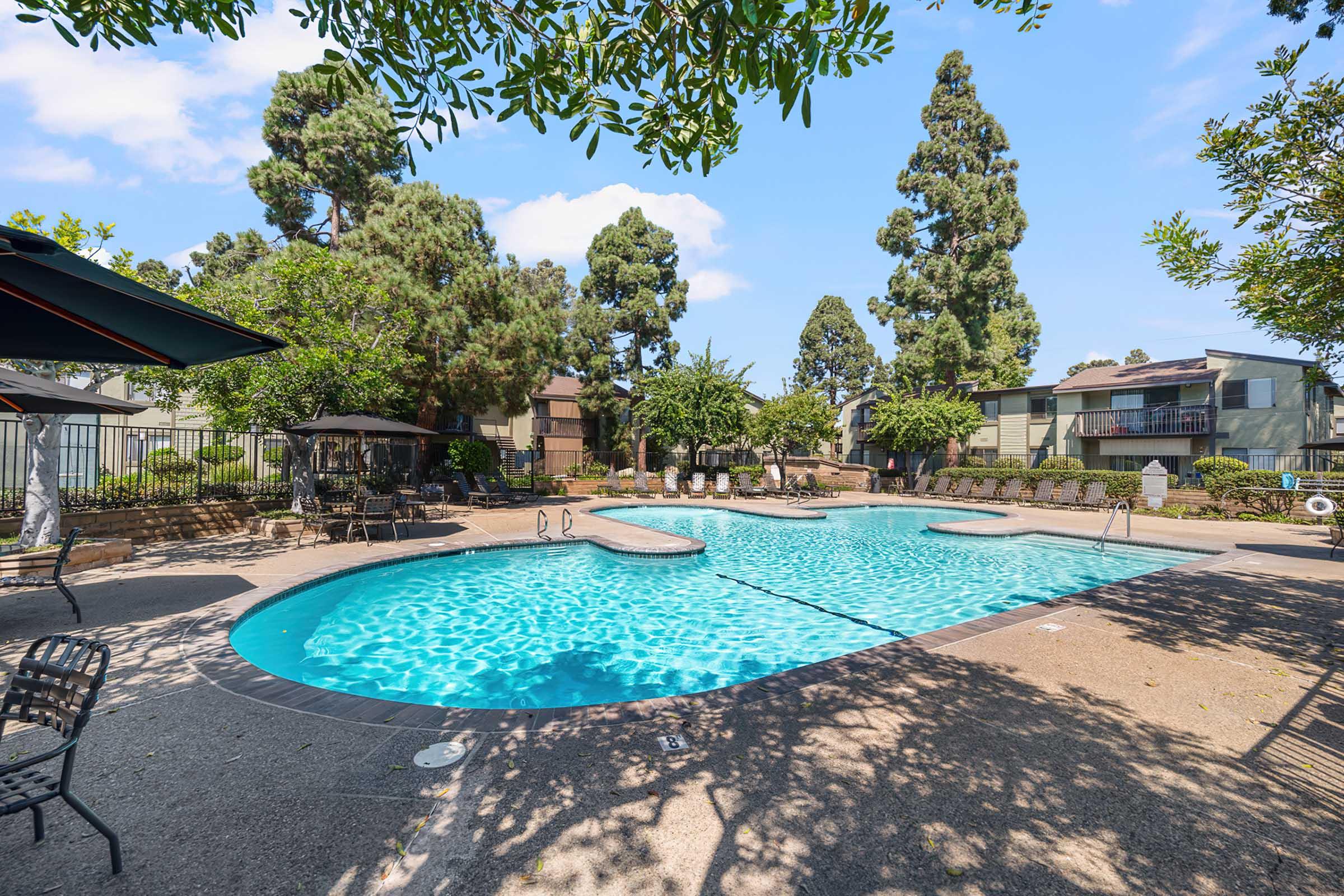 A serene outdoor swimming pool surrounded by lush green trees and landscape. The pool features a unique shape with steps leading in. Lounge chairs are placed around the pool area, and several apartments can be seen in the background, creating a relaxing residential atmosphere.