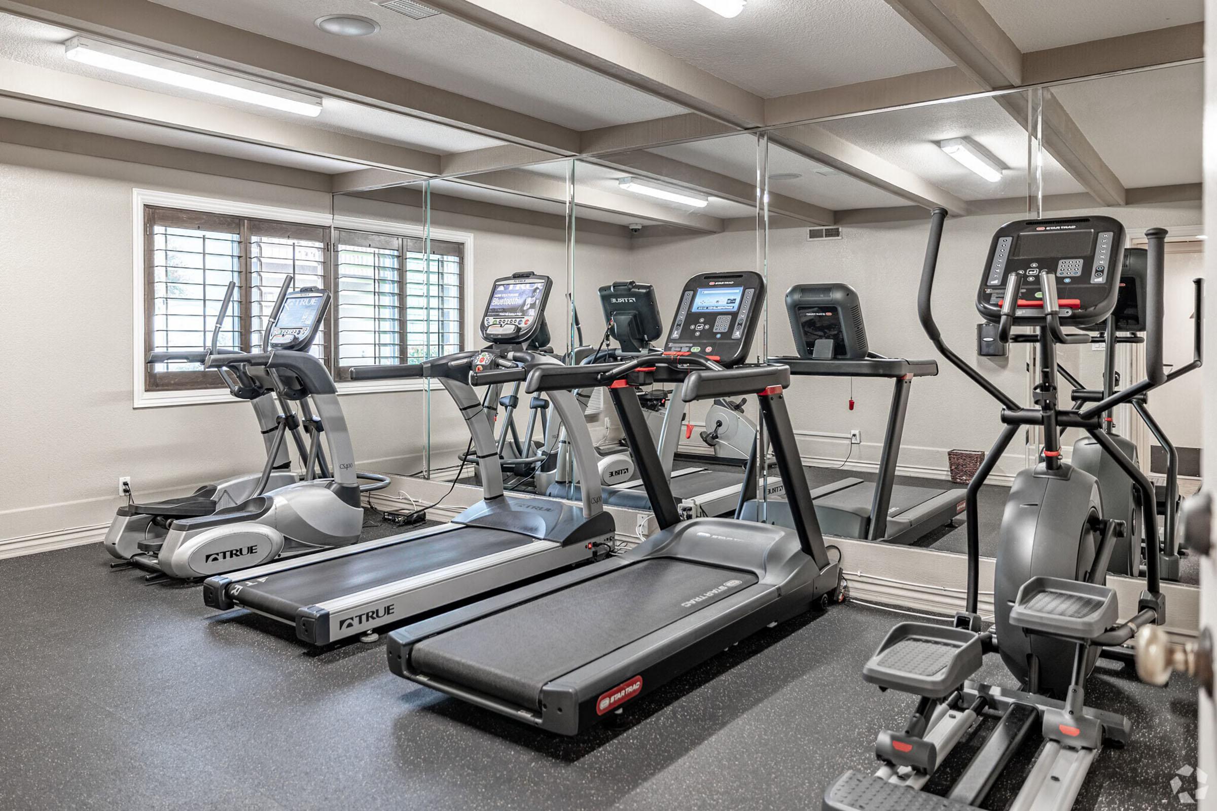 A modern fitness room featuring multiple treadmills, elliptical machines, and stationary bikes. The space is well-lit, with mirrors on one wall and a clean, black rubber floor surface, creating an inviting atmosphere for workouts.