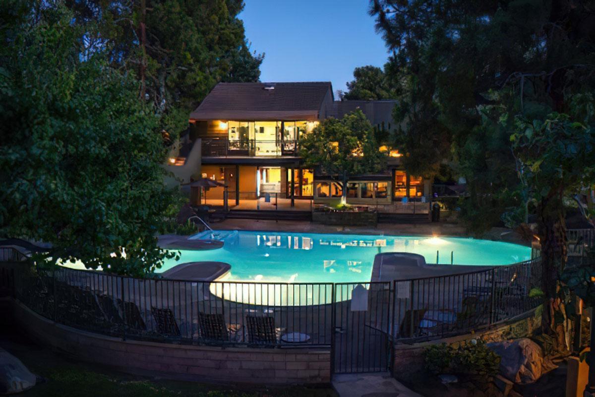 A serene evening scene featuring a brightly lit swimming pool surrounded by trees and lounge chairs. In the background, a modern building with large windows showcases warm interior lighting, creating a welcoming atmosphere. The pool water reflects the soft glow of the surrounding lights.